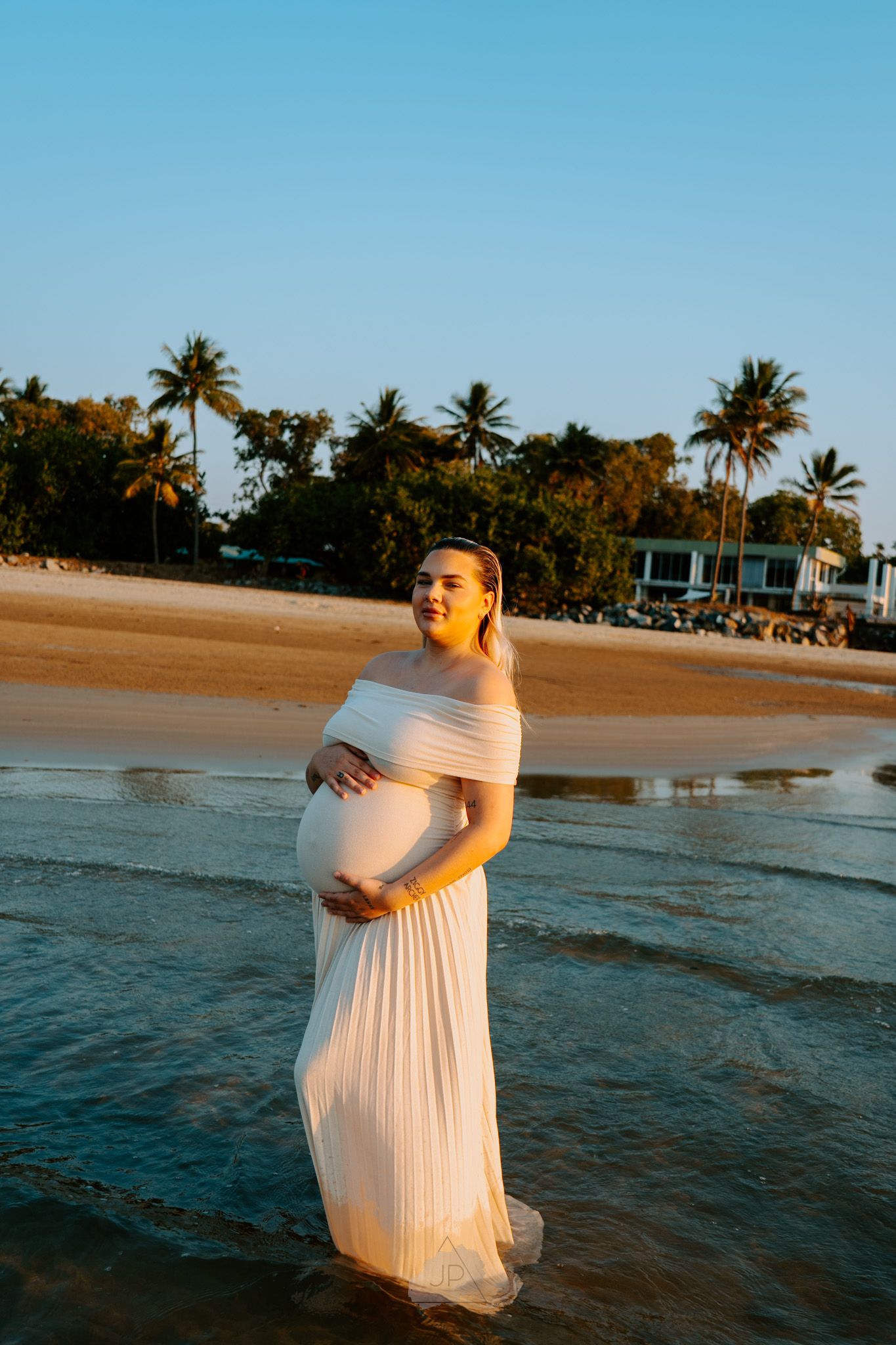 A person in a flowing white dress poses at sunset on a tropical beach with palm trees and ocean waves in view.