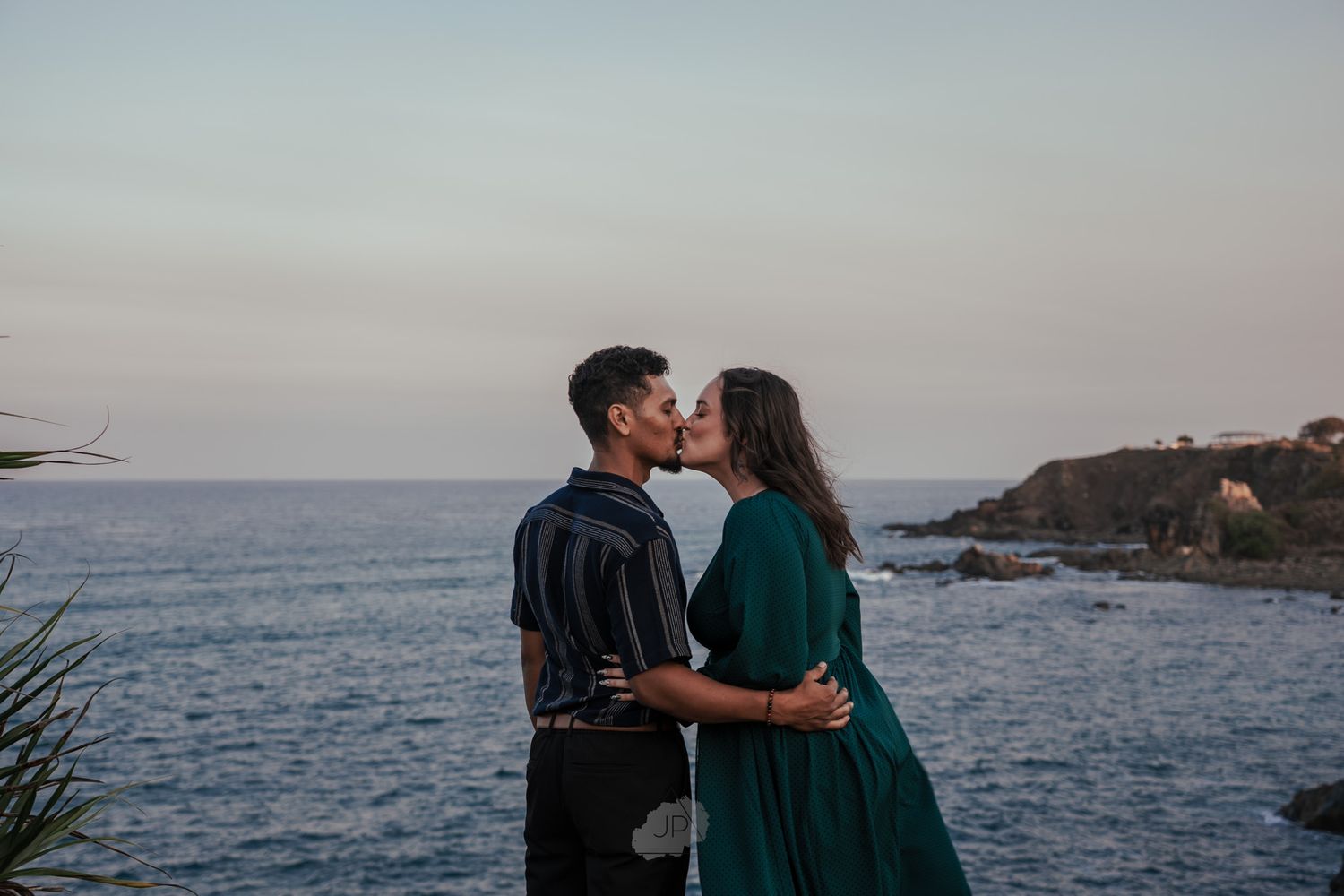 A romantic coastal scene at sunset with a couple embracing by the ocean with rocky cliffs in the background.