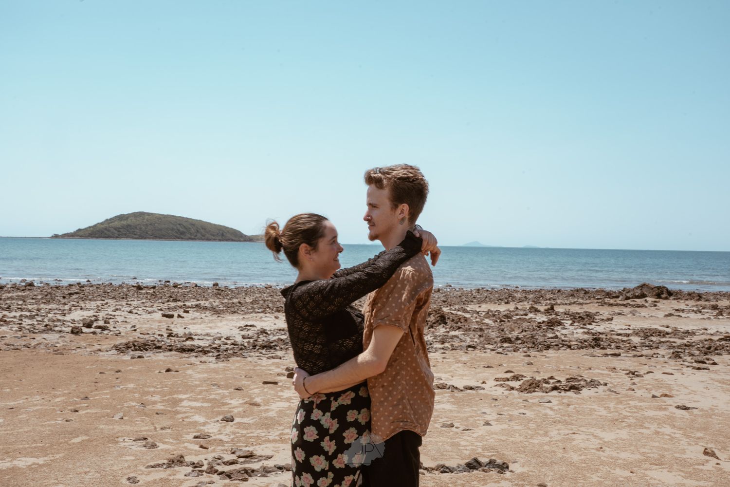 Couple embracing on a scenic rocky beach with ocean and mountain views in the background.
