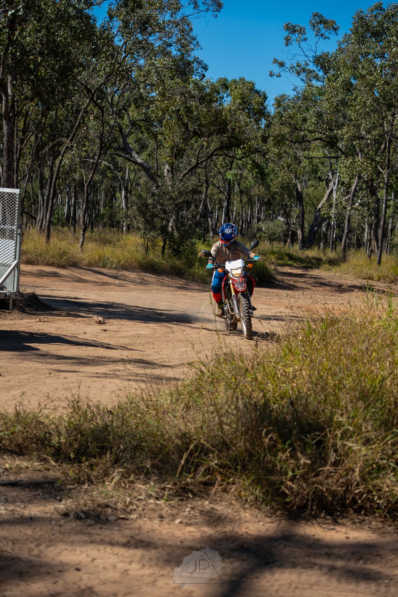 Dirt bike riders navigate a winding trail through Australian bushland under bright blue skies.