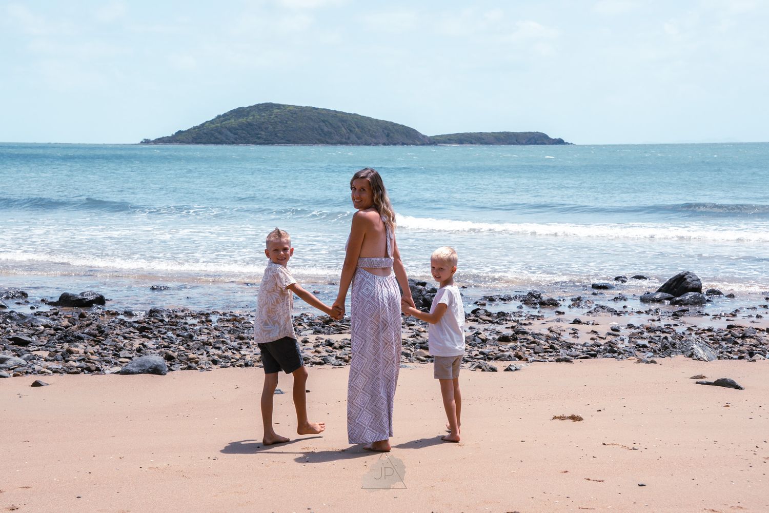 A family walks along a scenic beach shoreline with mountains visible in the distance on a sunny day.