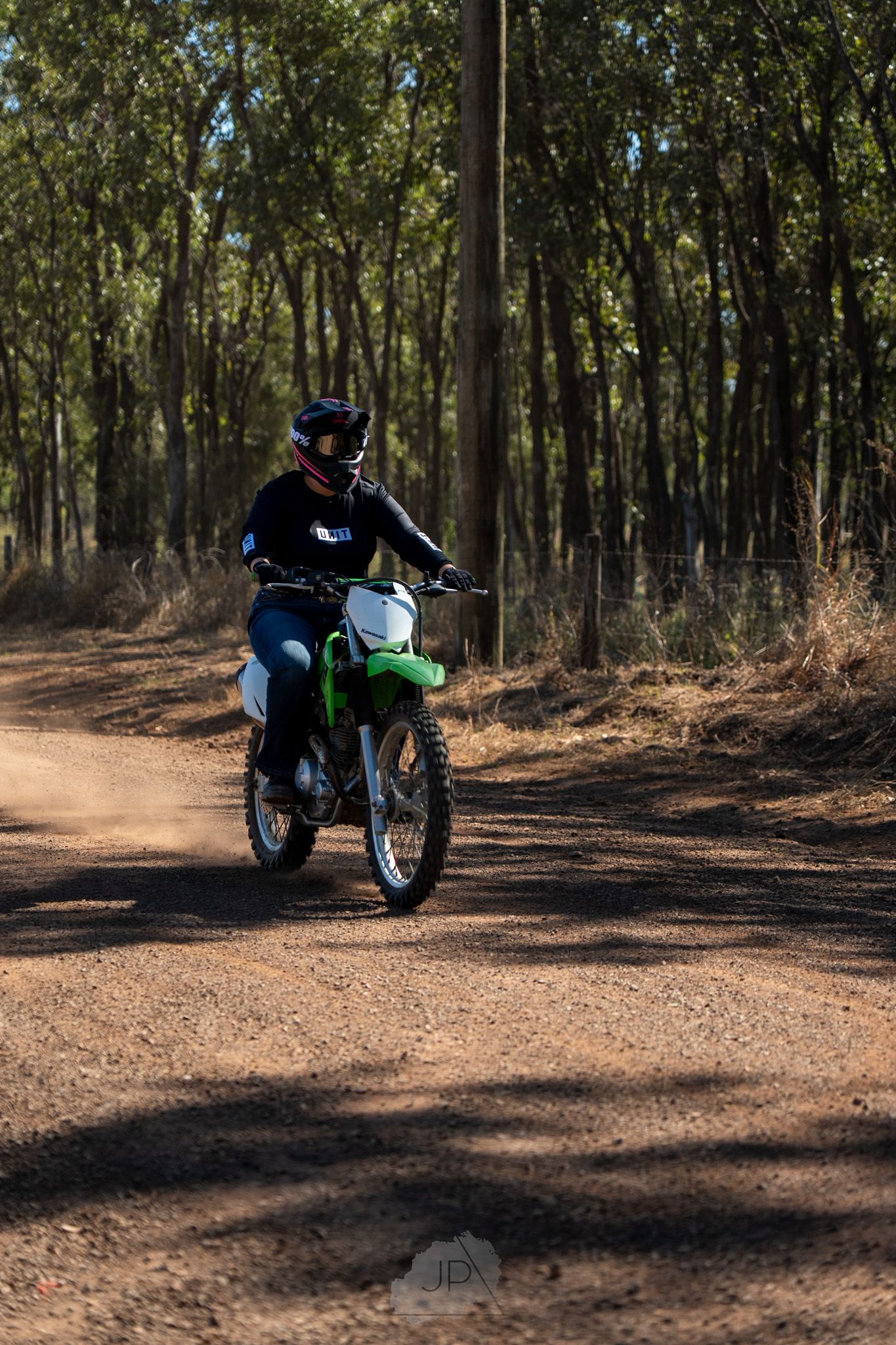 A dirt bike rider navigates through a wooded trail on a green off-road motorcycle in dappled sunlight.