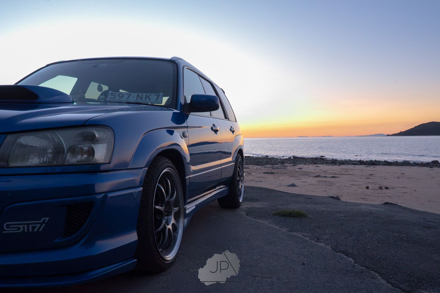 Classic blue Subaru WRX with lowered suspension cruising along scenic beachfront road at sunset.