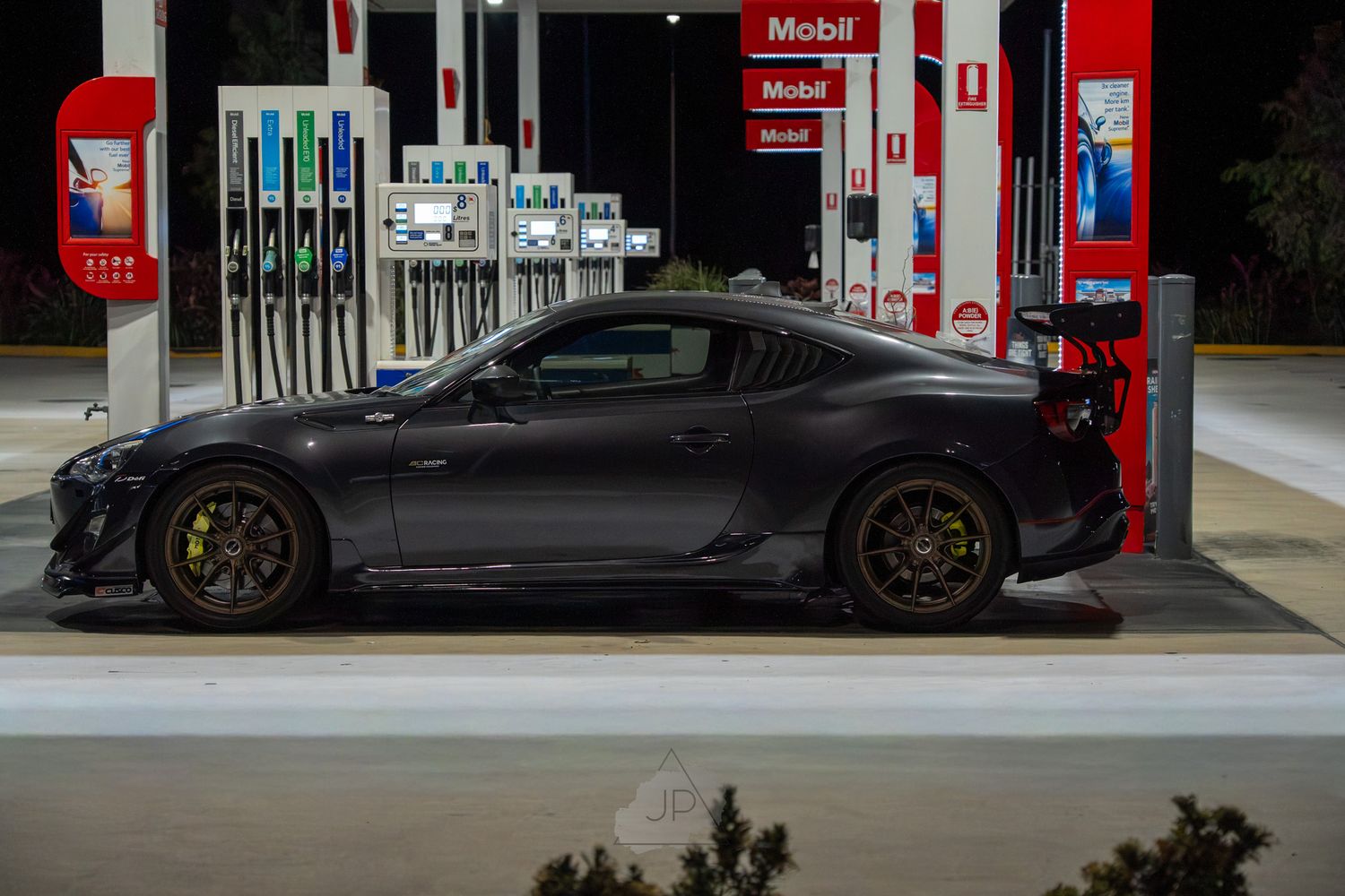Black sports car refueling at a Mobil gas station with multiple fuel pumps visible in the background.