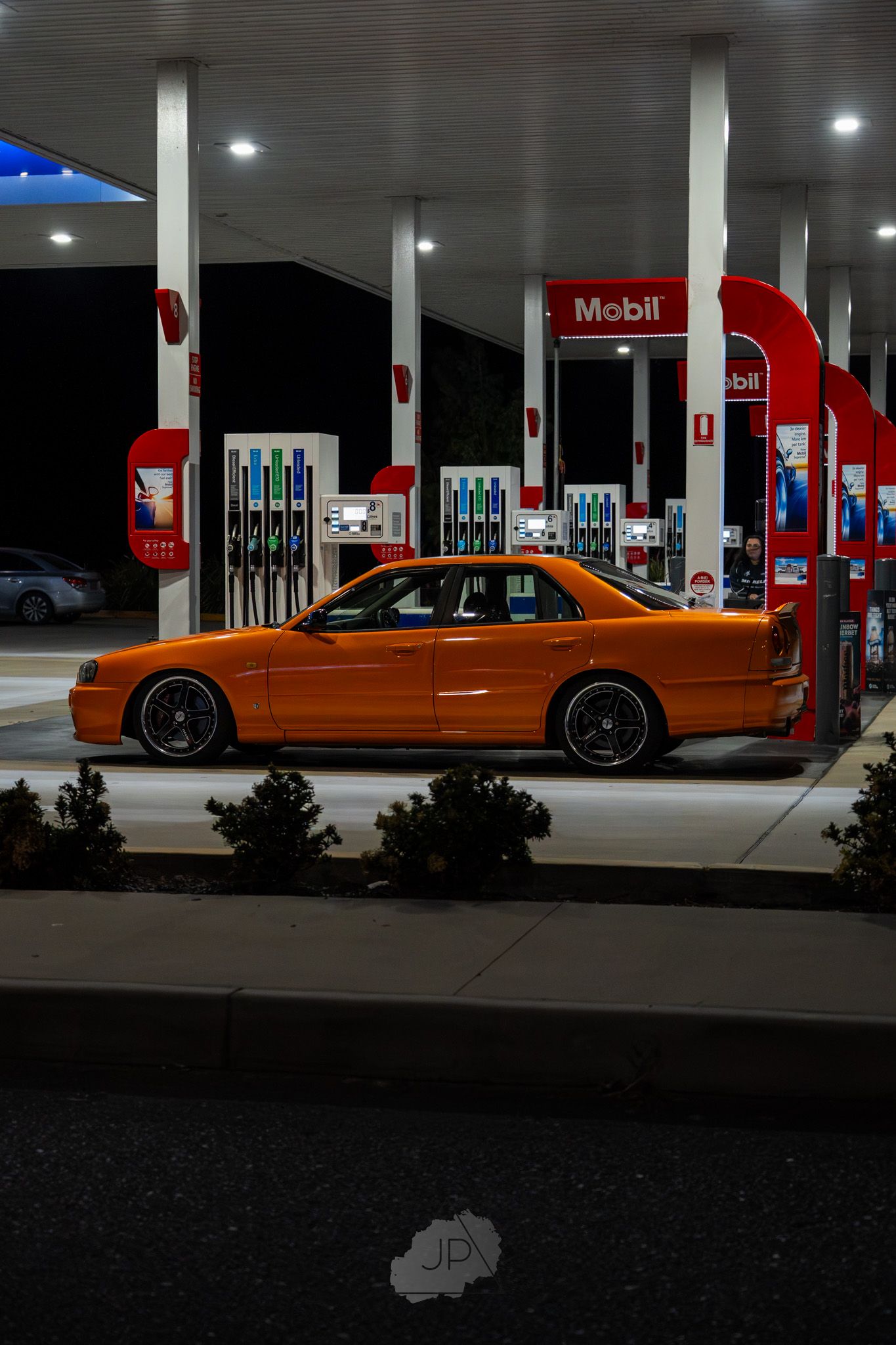 An orange sports car parked at a Mobil gas station at night with illuminated fuel pumps and canopy lights.