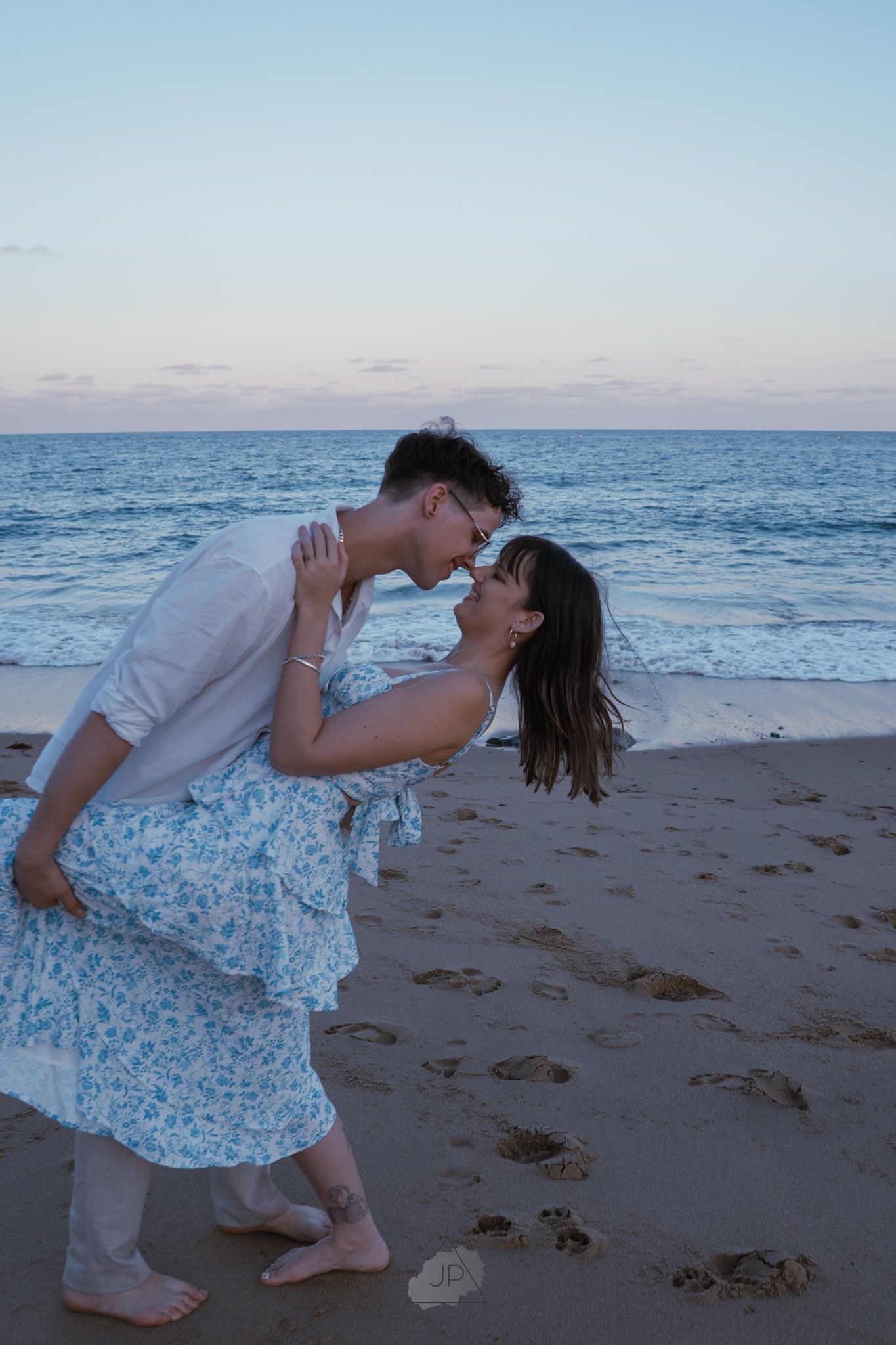 A romantic couple shares a tender kiss on the beach at sunset with ocean waves in the background.