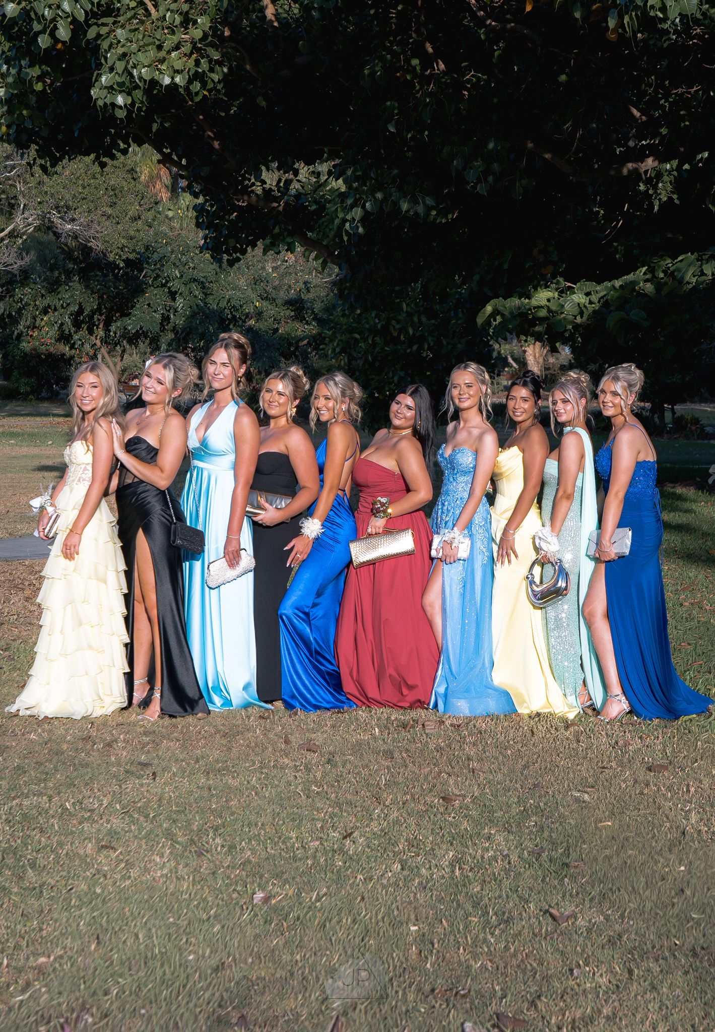 A group in formal evening gowns poses together outdoors in shades of blue, red and cream for a special occasion photo.