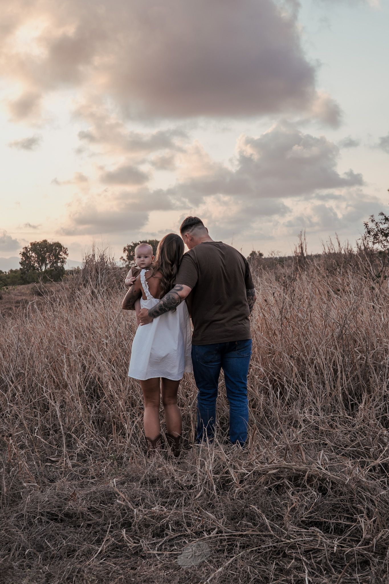 A couple stands together in a field of dried grass during sunset creating a romantic silhouette against the sky.