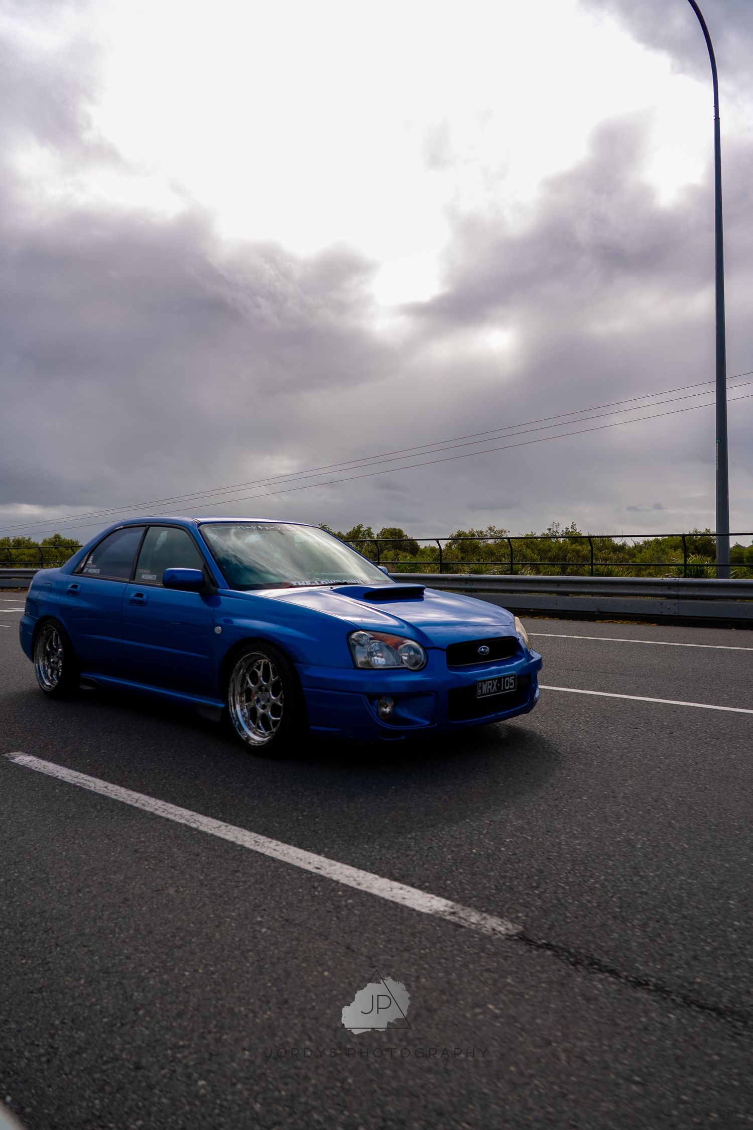 A blue Subaru WRX STi with aftermarket modifications parked on a highway under dramatic cloudy skies.