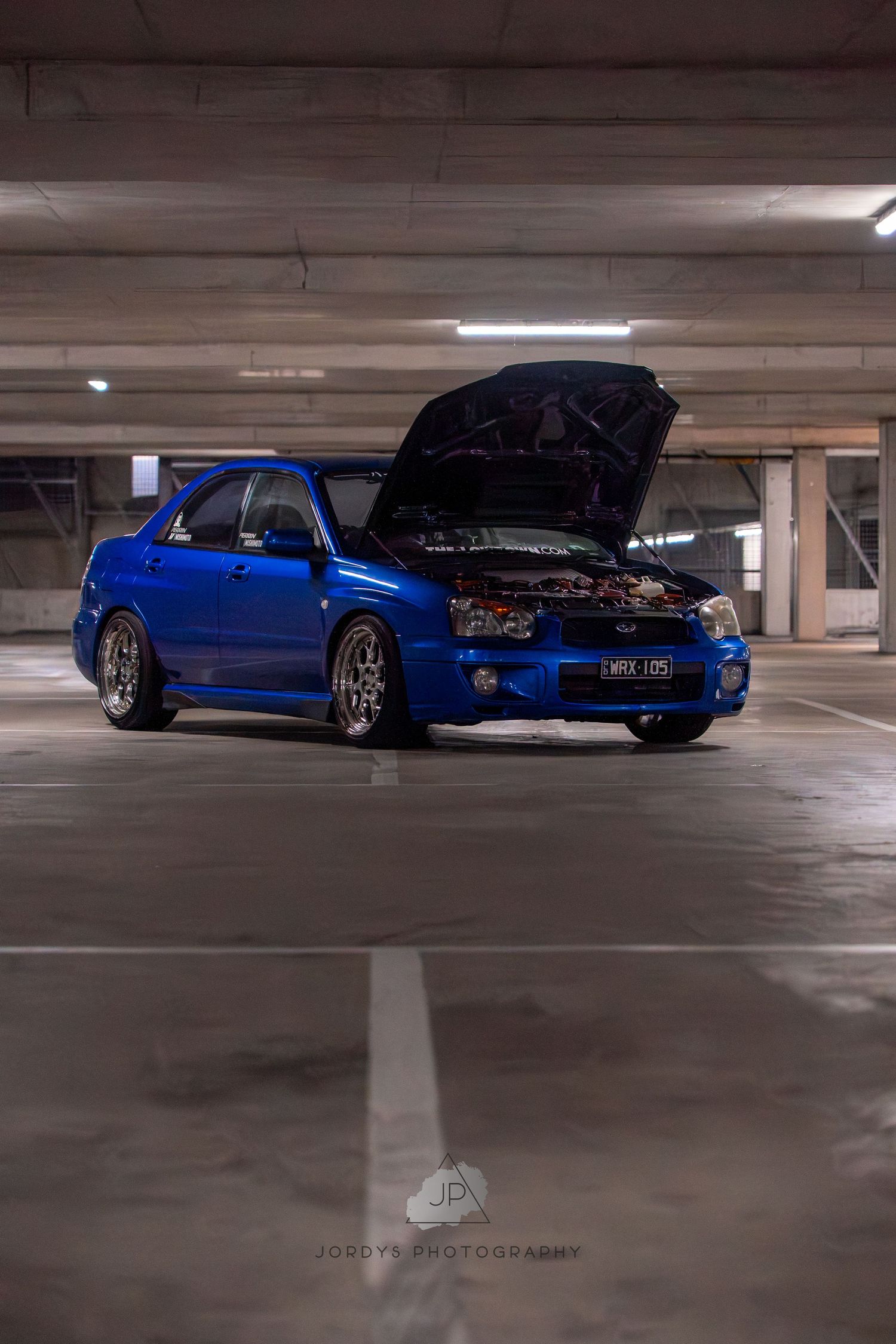 Blue modified sports car with open hood parked in dimly lit concrete parking garage.