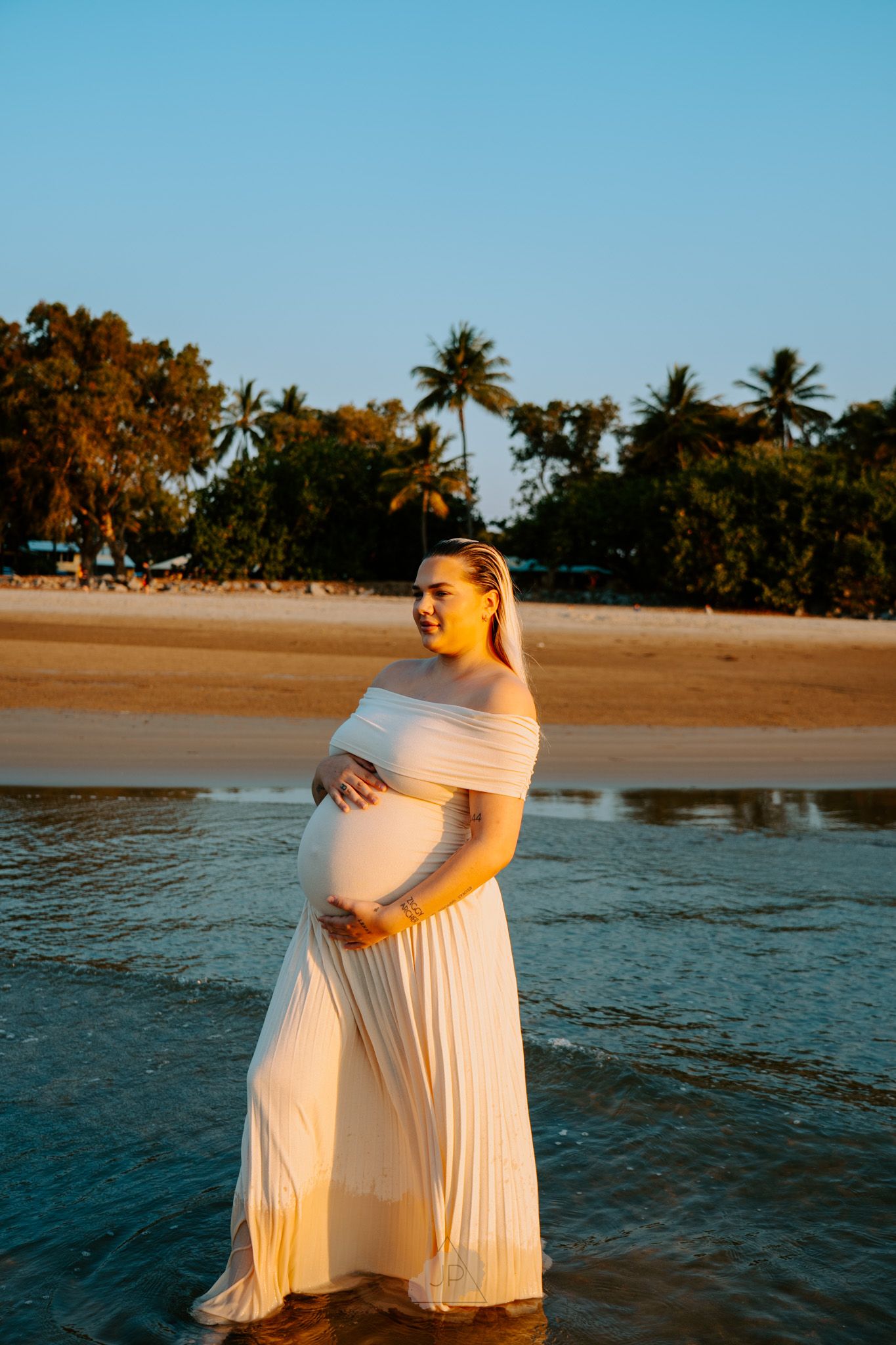 A person in a flowing white dress poses at sunset on a tropical beach with palm trees in the background.