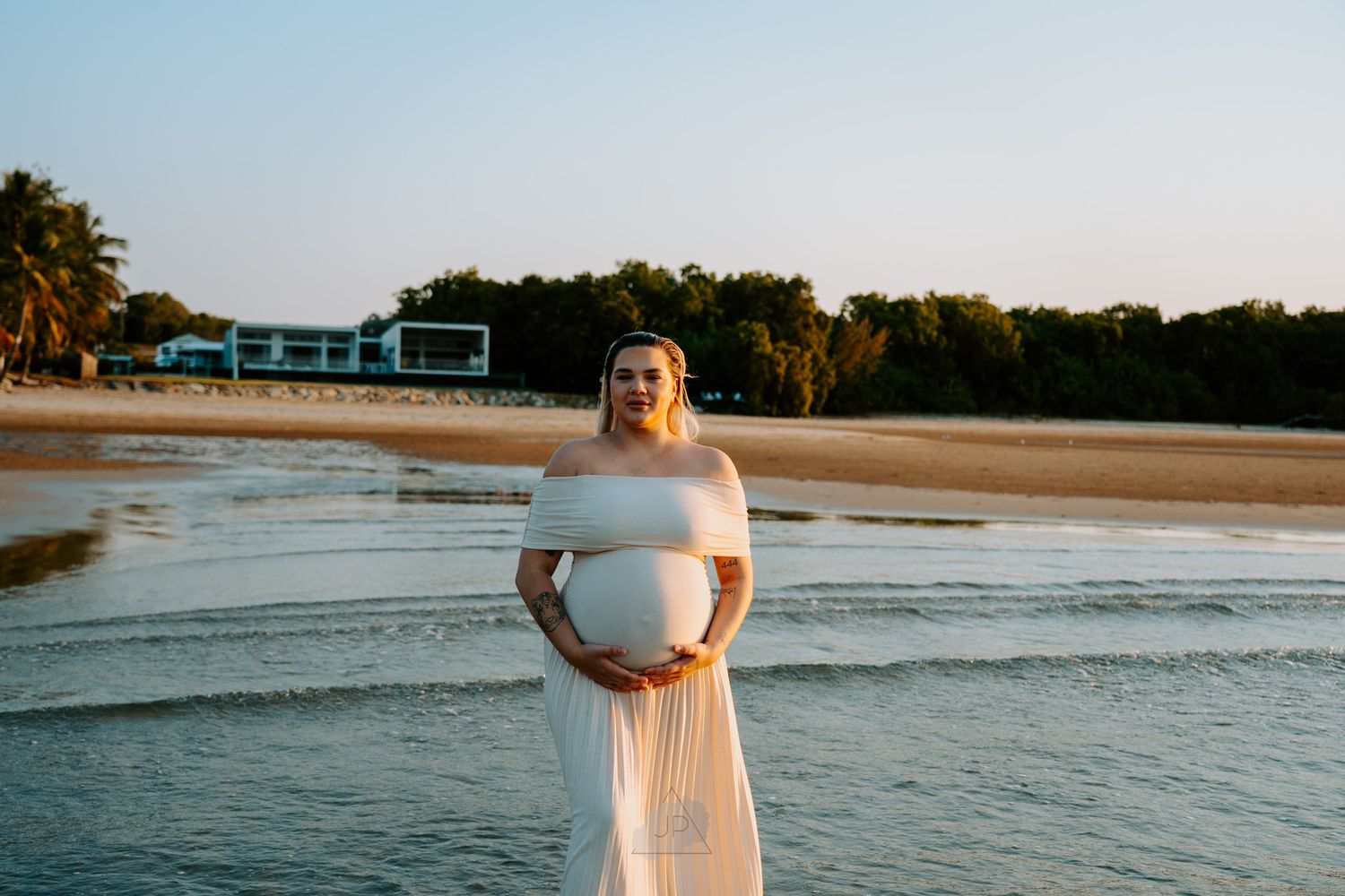 A person in a white dress walks along a sandy beach at sunset with ocean waves rolling in.