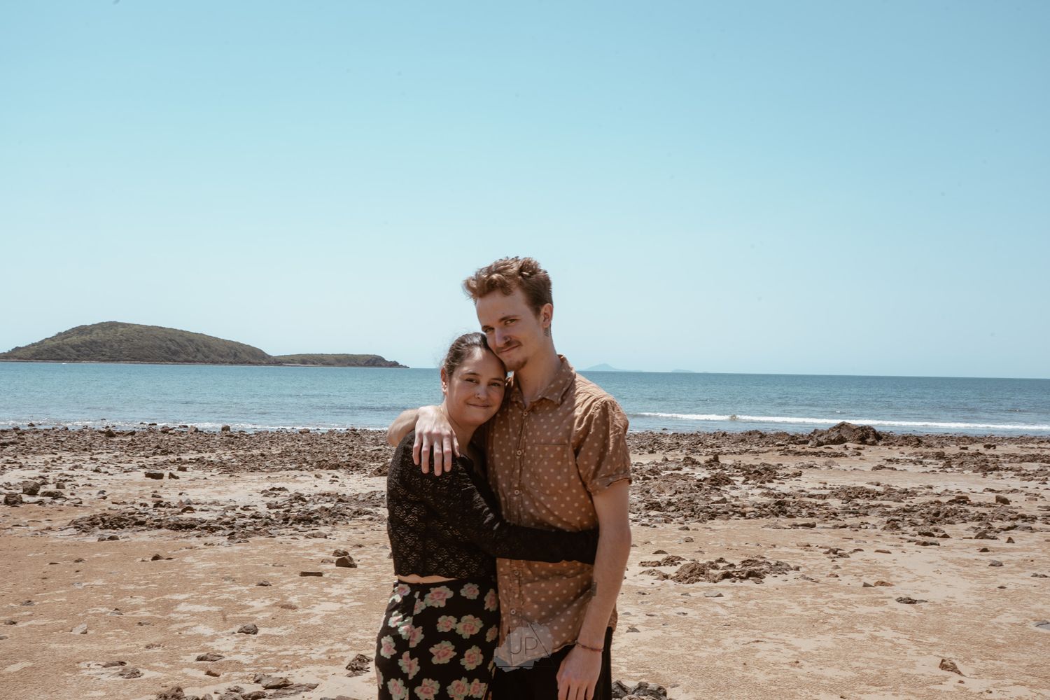 A couple embraces on a rocky beach with ocean waves and blue sky in the background.