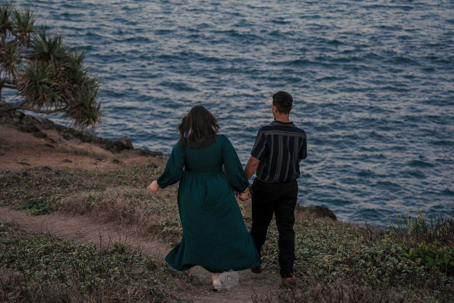 Two people in casual attire stroll together on a grassy oceanside trail during sunset.