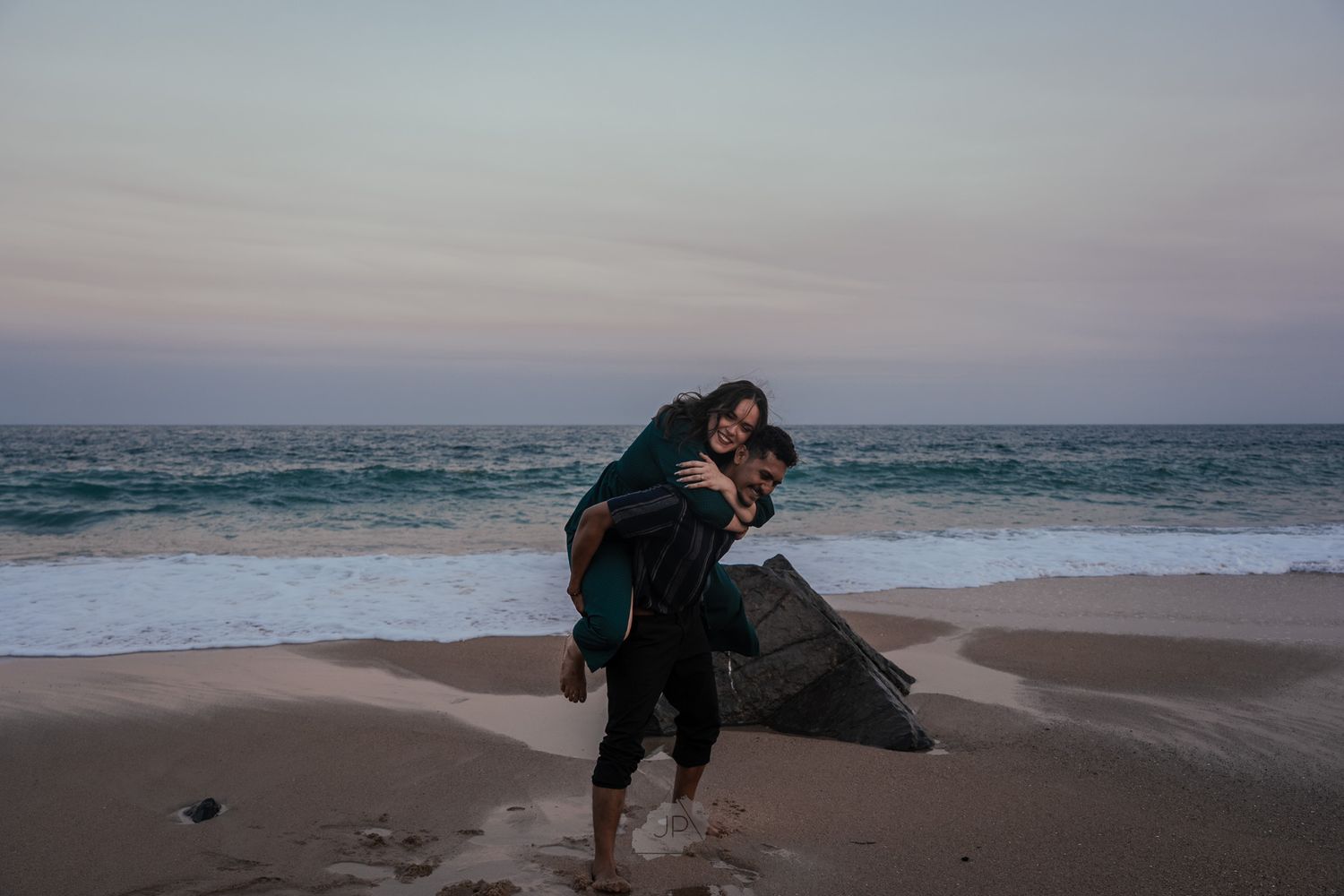 A couple embraces on a sandy beach at dusk with ocean waves and cloudy sky in the background.