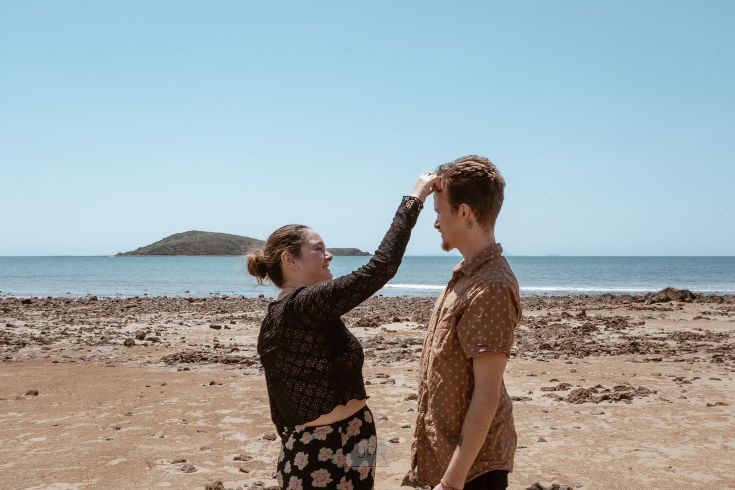 Two people interact on a sunny beach with mountains and ocean in the background.