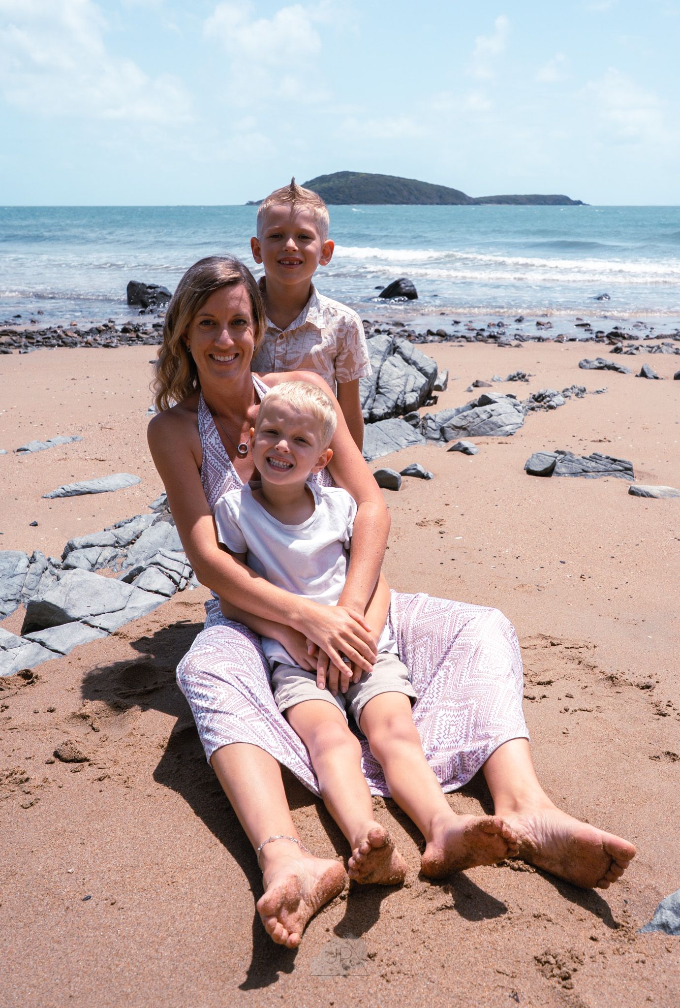 Two people sitting together on a sandy beach with rocks and ocean views in the background.