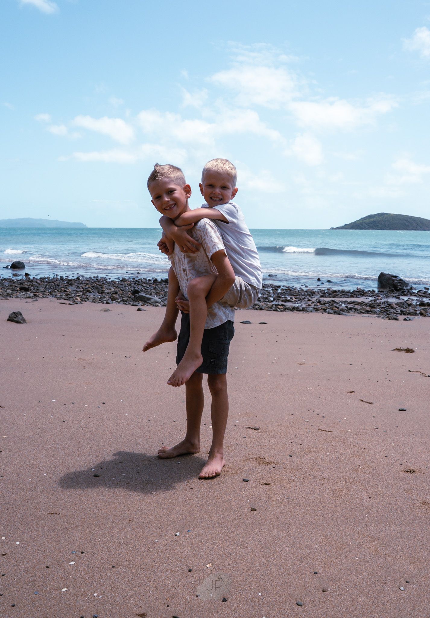 Two people playfully interacting on a sandy beach with ocean waves and rocky coastline in the background.
