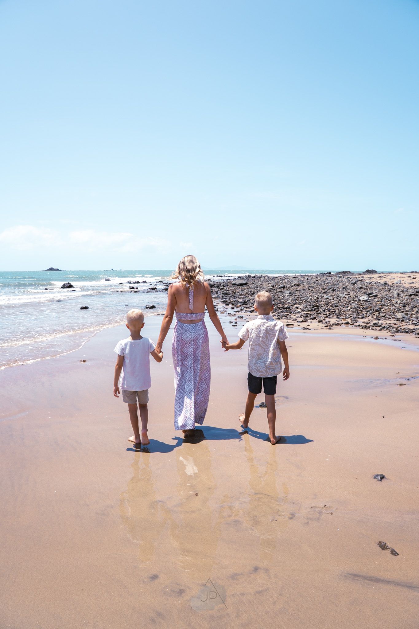 Three people walking hand in hand along a sunny beach with rocky coastline and blue ocean in the background.