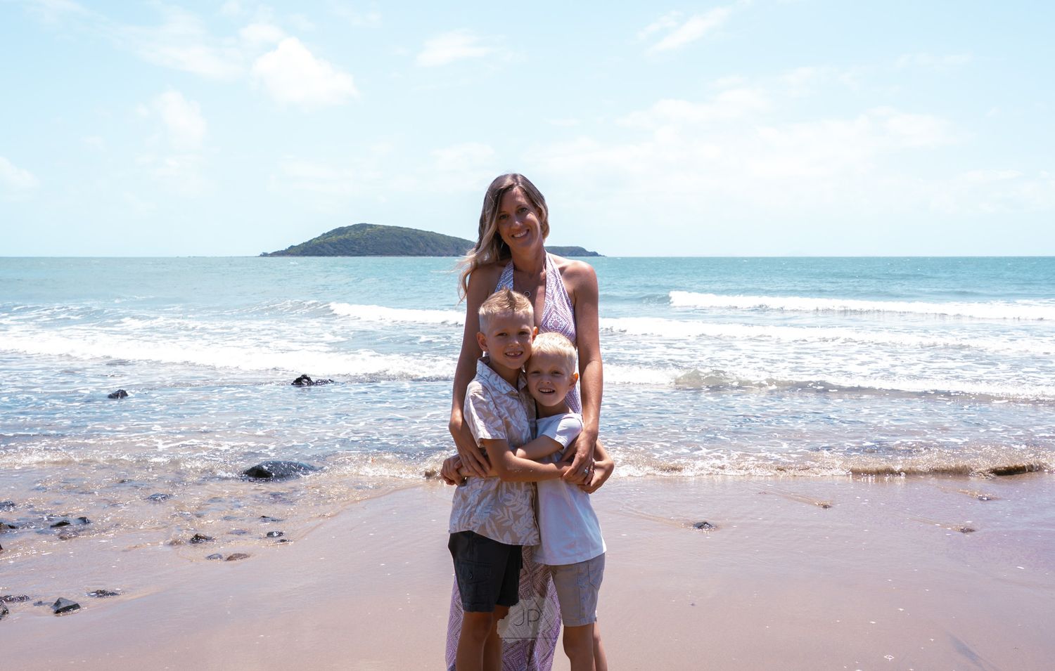 A couple embraces on a sunny beach with ocean waves and mountains visible in the background.