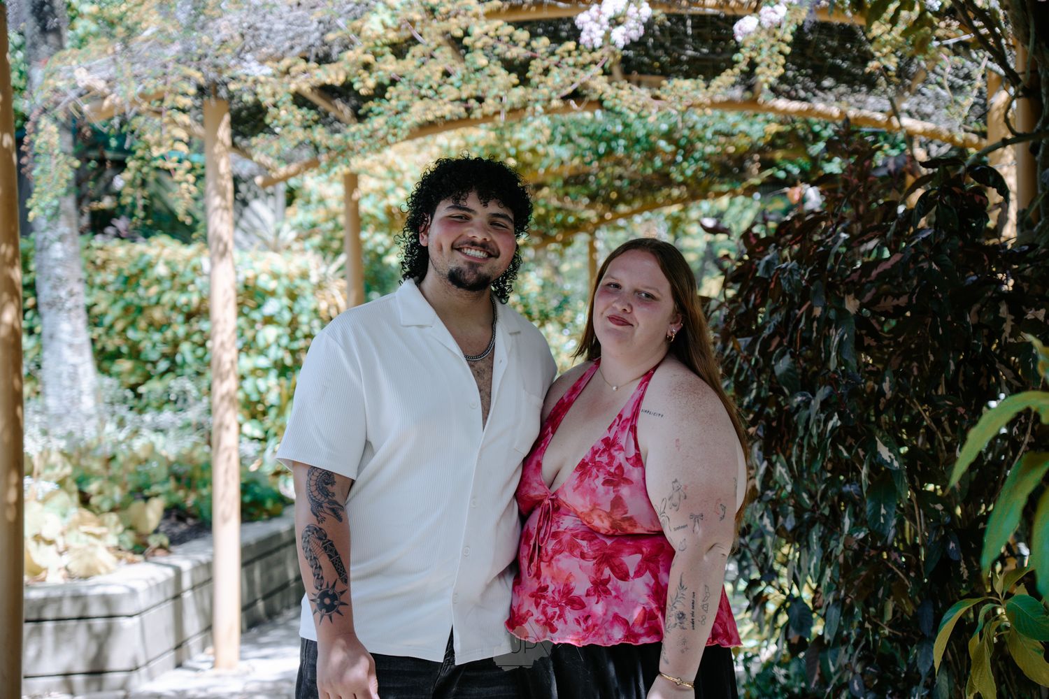 A couple poses together outdoors on a sunny day surrounded by lush greenery and trees.