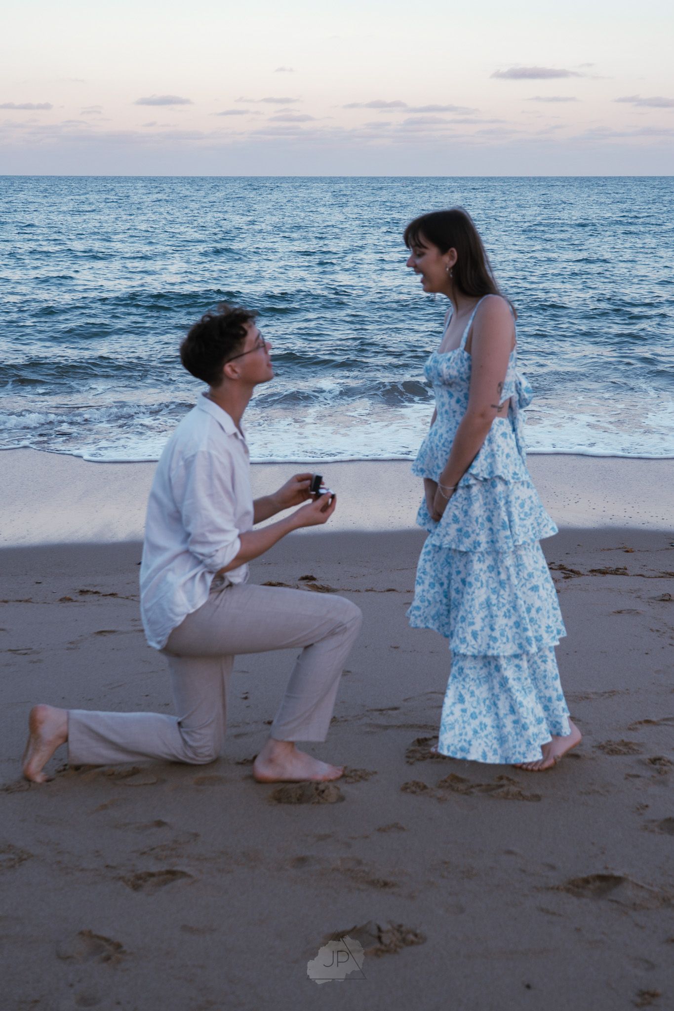 A romantic beach proposal scene at sunset with a couple in white and blue attire by the ocean waves.