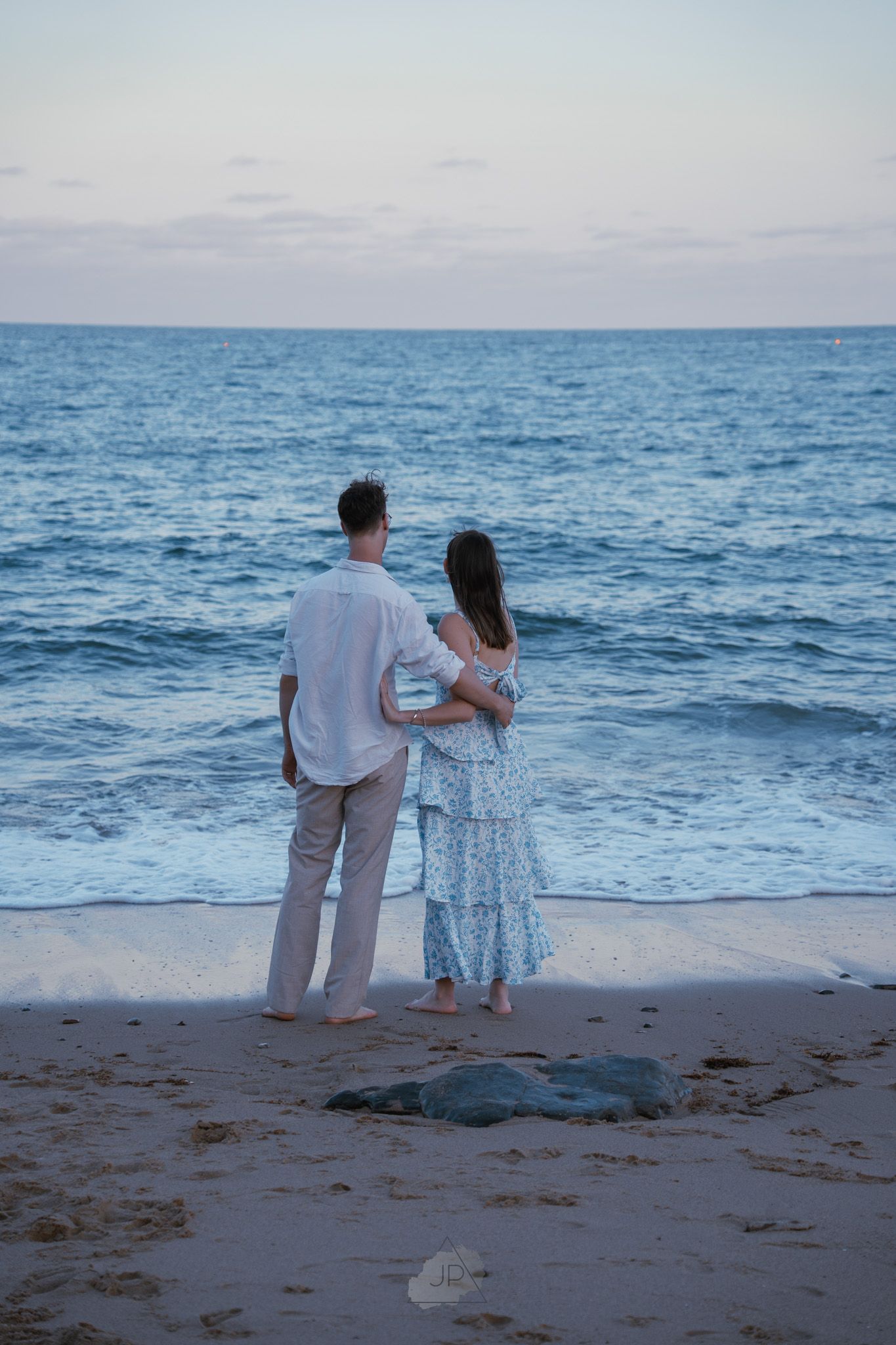 A couple stands together on a beach at dusk overlooking the blue ocean waves.