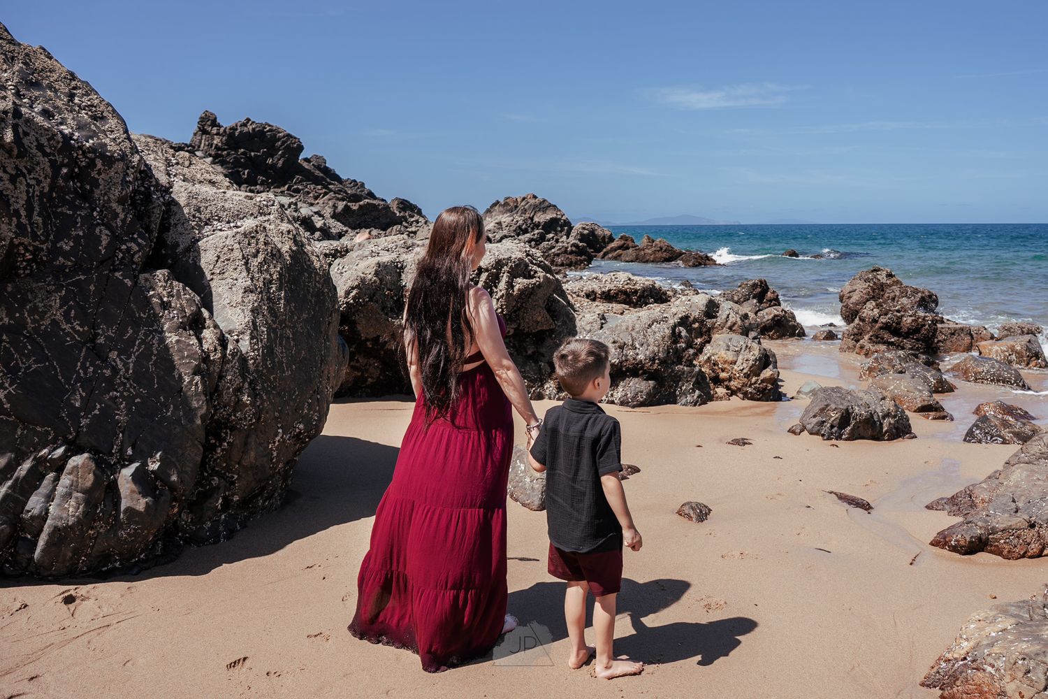 Two people explore a rocky beach coastline with large boulders and turquoise ocean waters on a sunny day.