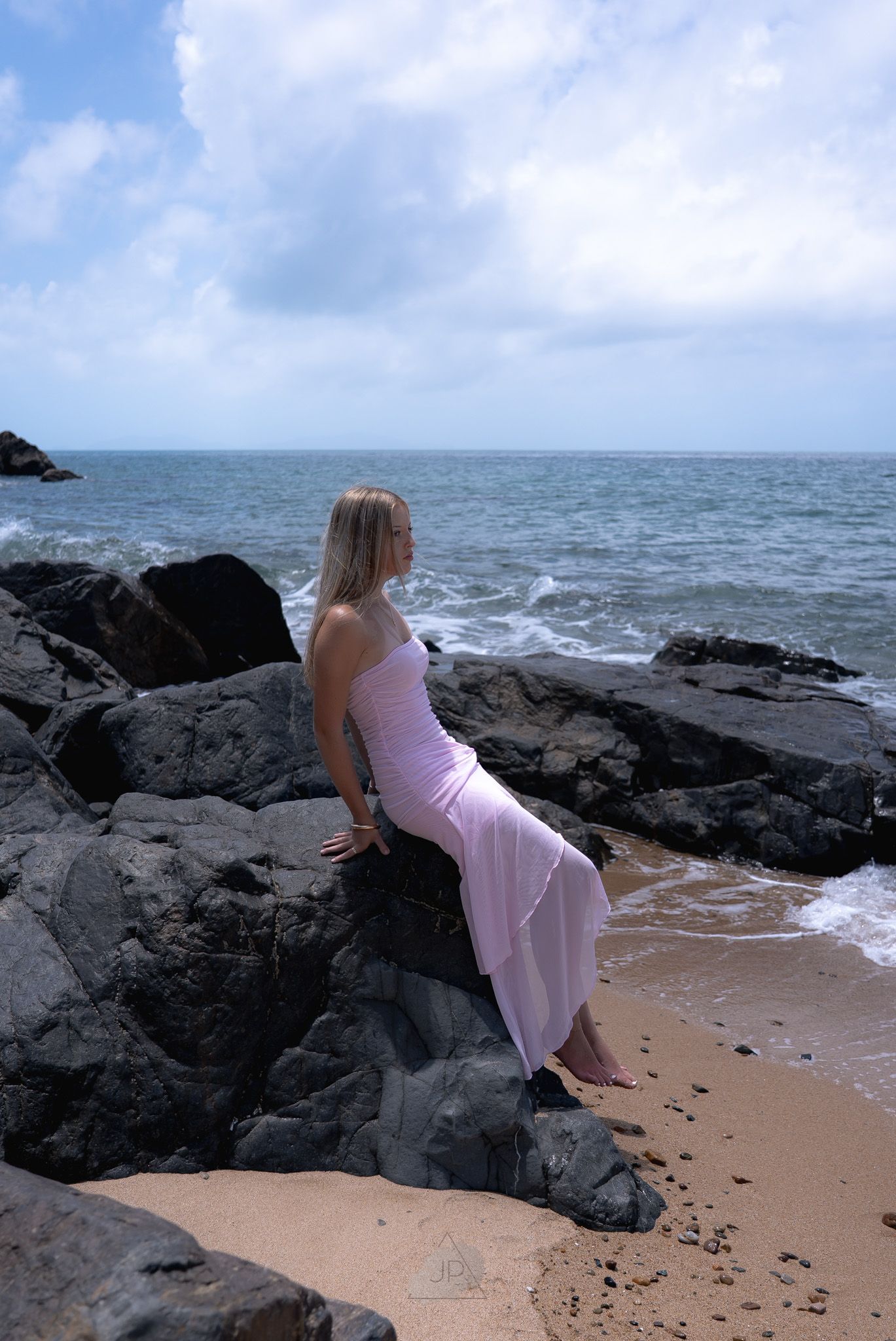 A person in a pink dress lies on dark volcanic rocks along a sandy beach with ocean waves in the background.