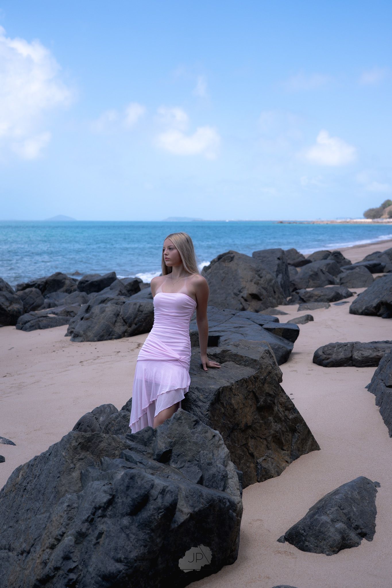 A person in a pink dress sits on dark volcanic rocks along a tropical beach with turquoise ocean waters in the background.