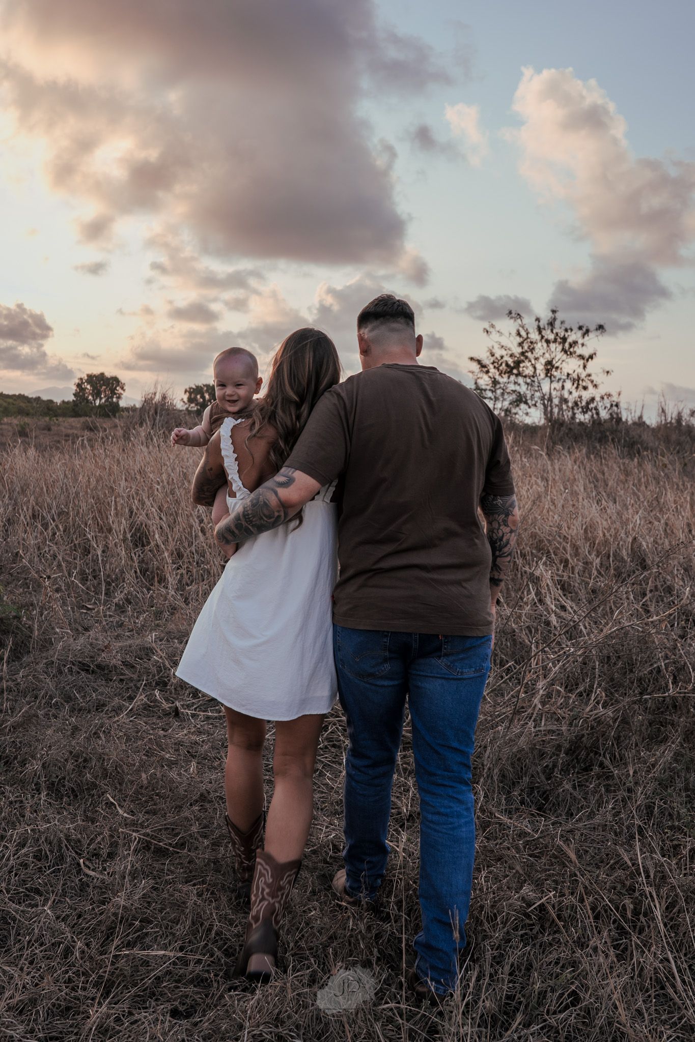 A couple and their baby stand together in a brown field at sunset with cloudy skies above.