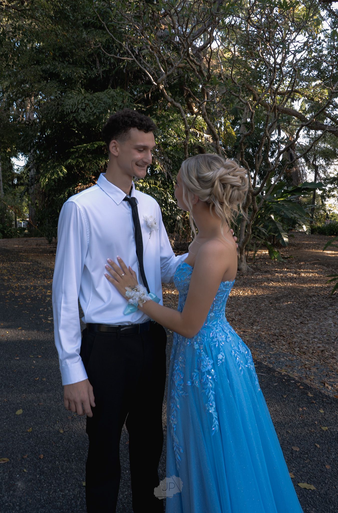 A couple in formal attire, with a blue ballgown and white dress shirt, share a romantic moment outdoors.
