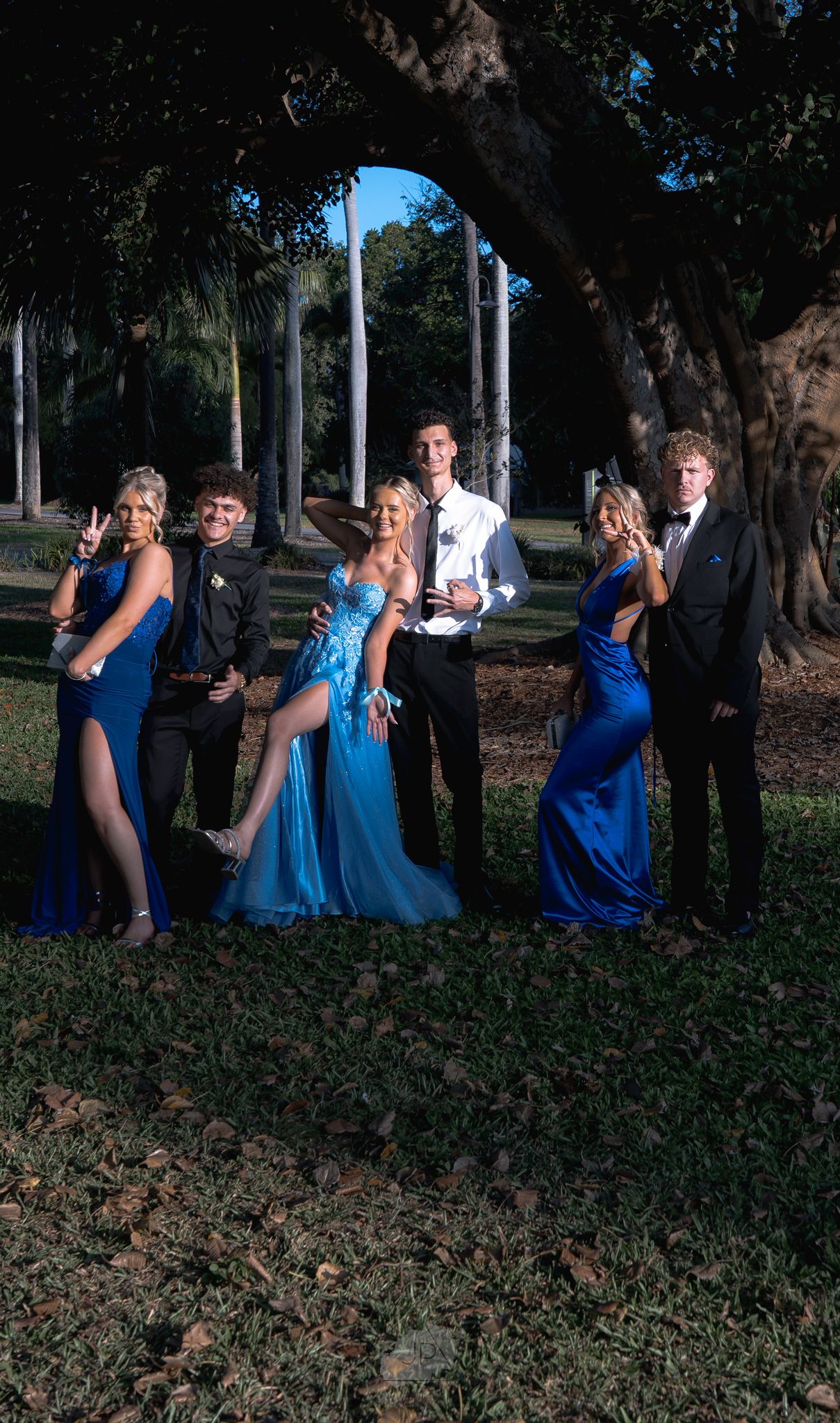 Wedding party poses playfully in a row wearing coordinated blue dresses and formal attire in an outdoor garden setting.