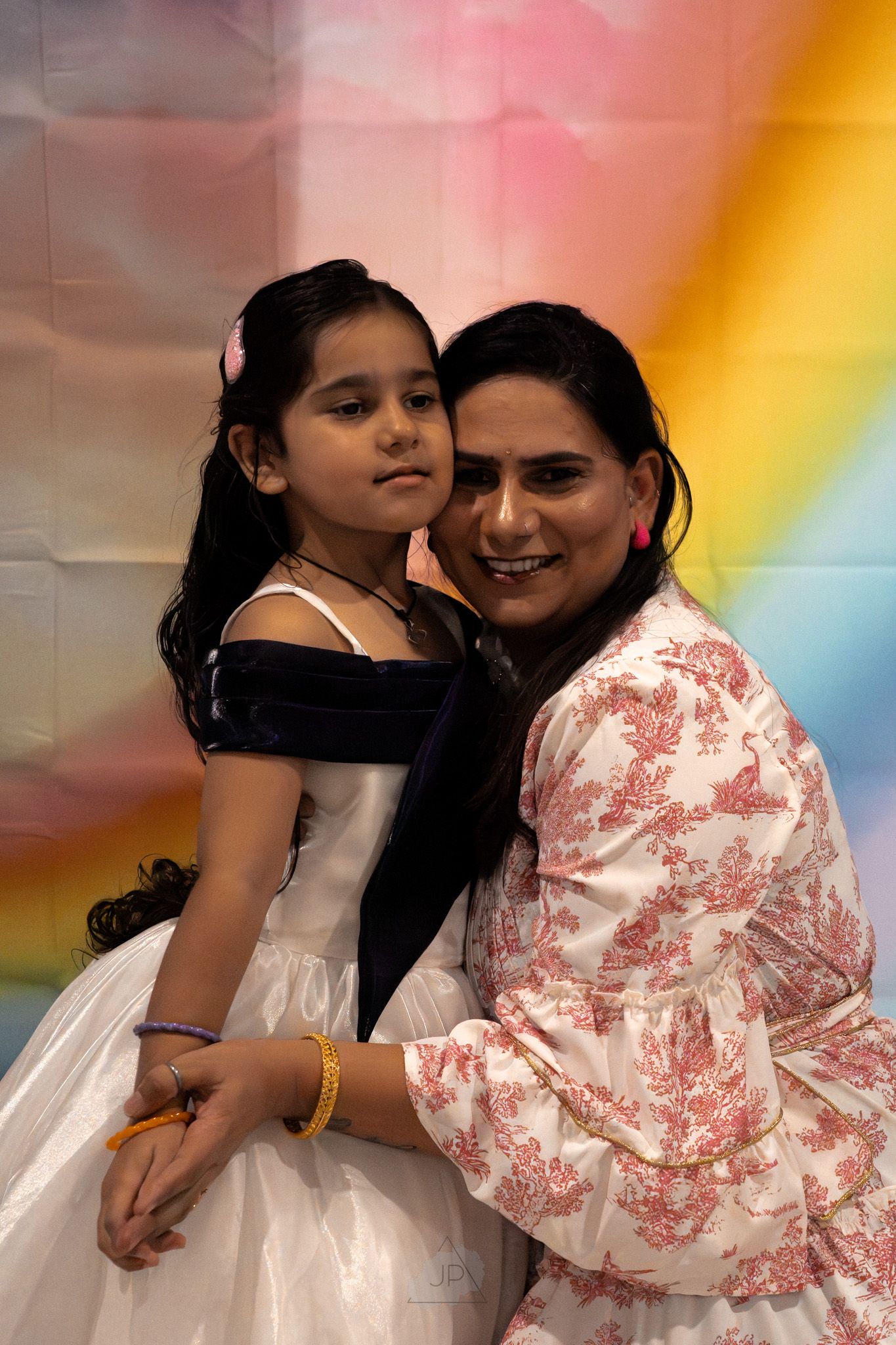 A child in a white dress poses with an adult wearing a pink and white patterned top against a colorful rainbow backdrop.
