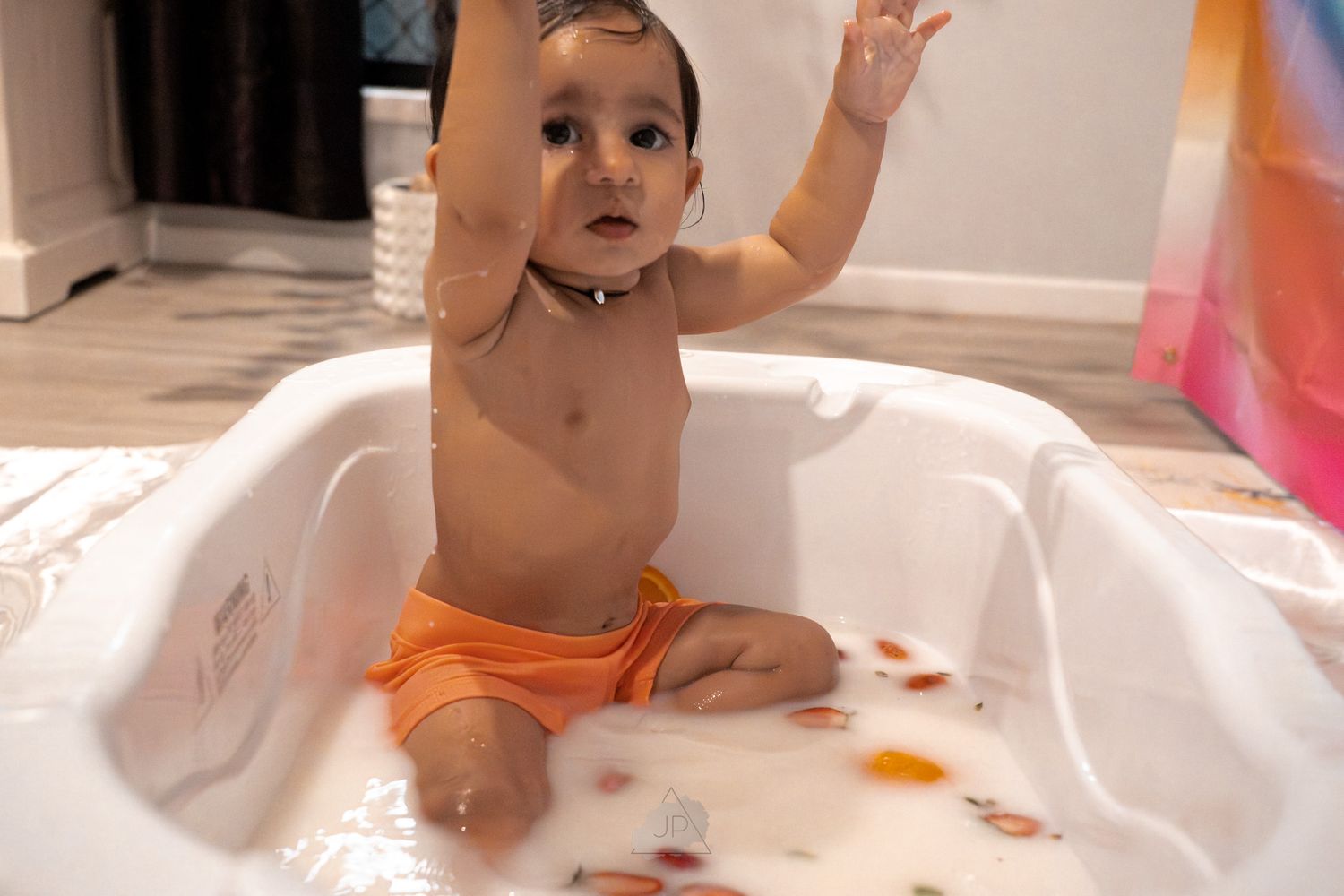 Baby in orange diaper sits in white bathtub playing with colorful bath toys during bathtime.