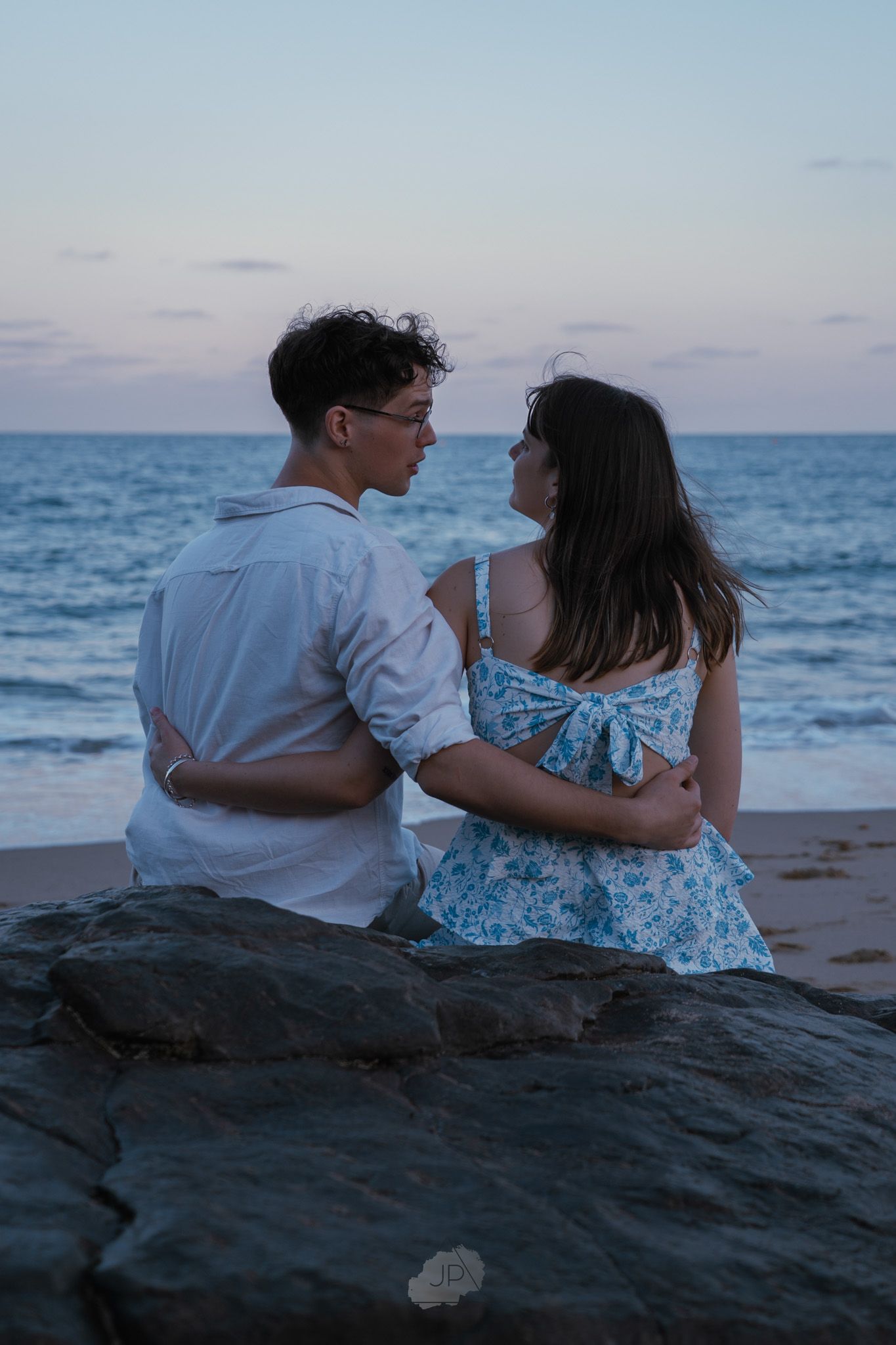 A couple sits together on coastal rocks watching the sunset over the ocean in matching white and blue outfits.