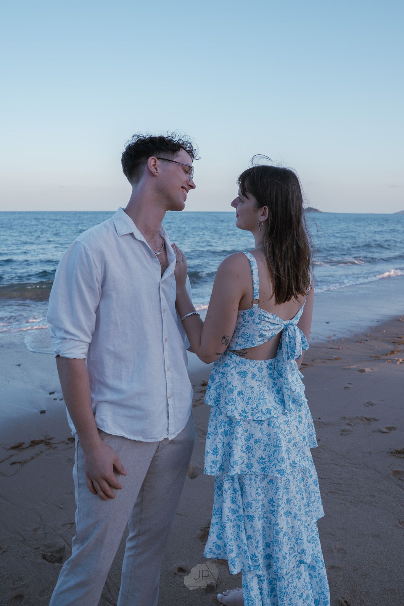 A couple shares a romantic moment together on a beach at sunset wearing white and blue floral outfits.