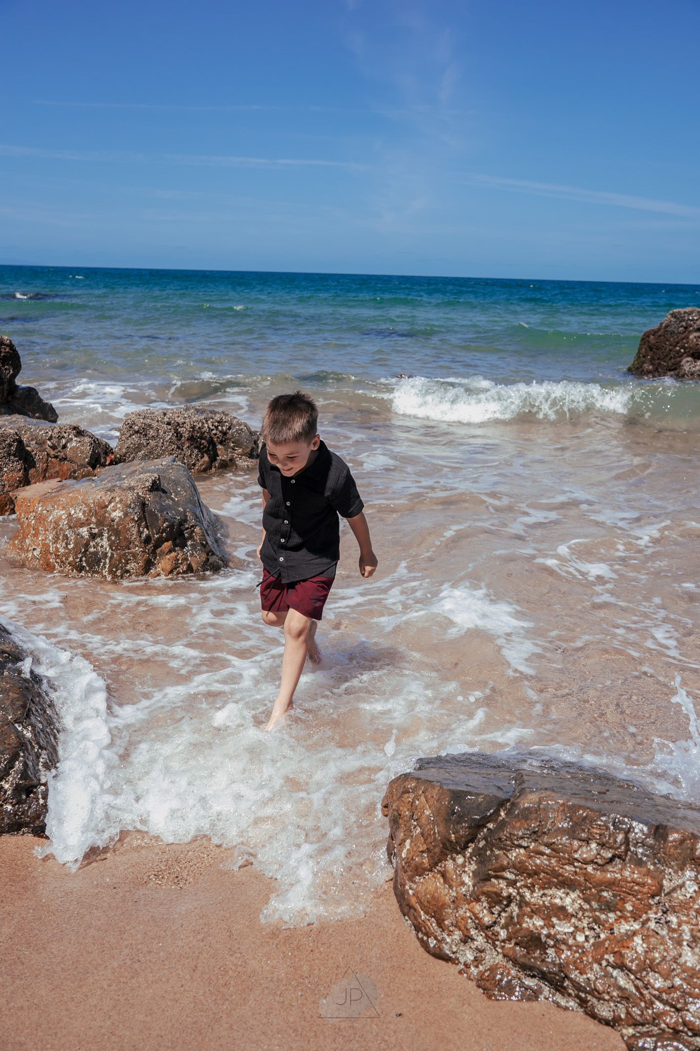 A child plays in the ocean waves crashing against rocky outcrops on a sunny beach with turquoise waters.