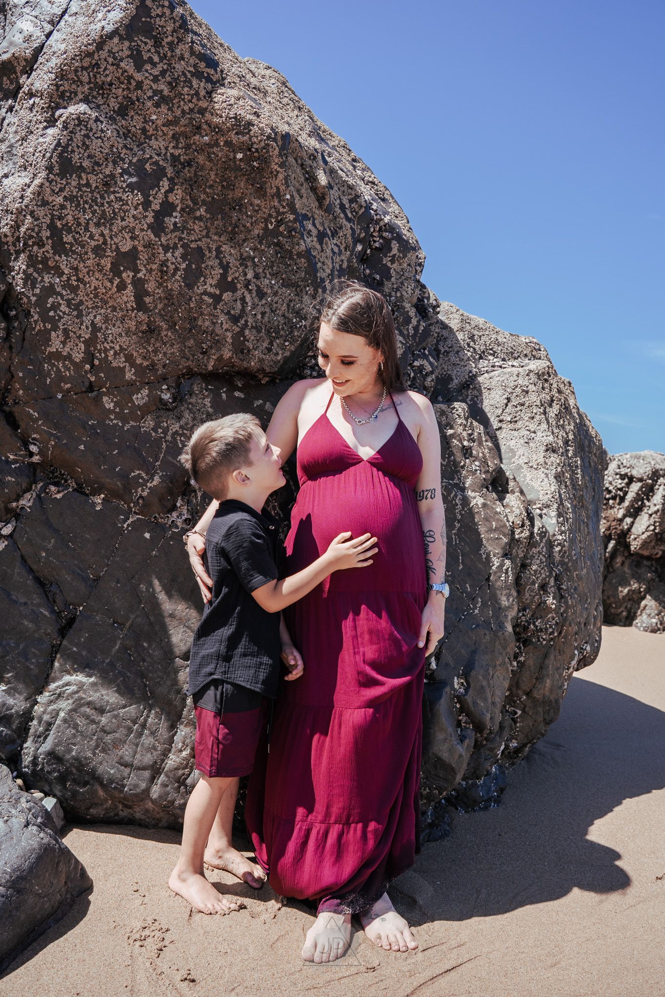 A couple shares a tender moment against a rocky beach backdrop in a maternity photo sequence.