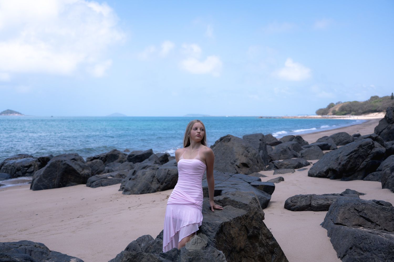 Person in pink dress walking along scenic rocky beach coastline with blue ocean and sky in background.