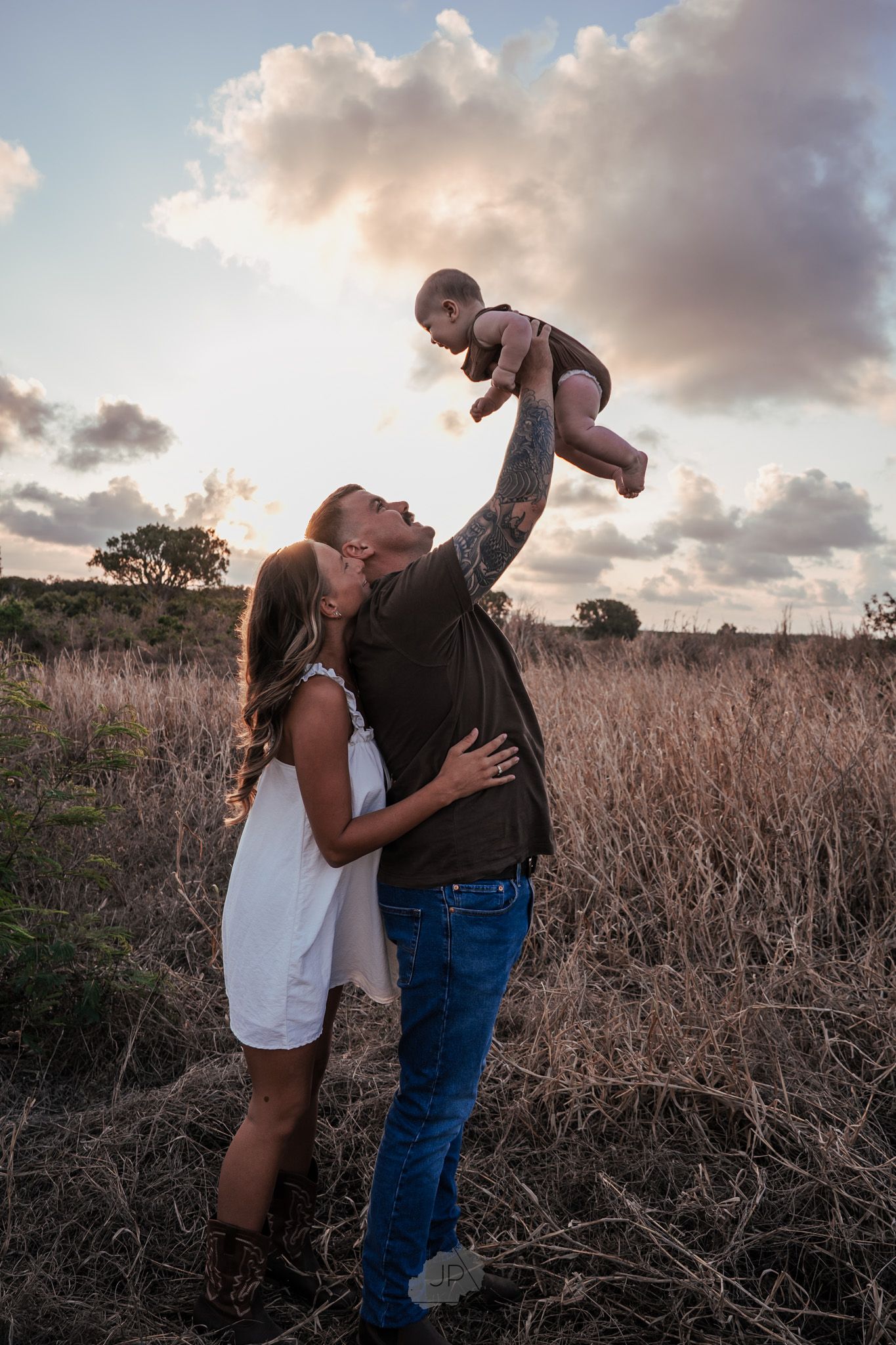 A couple dances playfully in a field of dried grass and wildflowers during sunset.