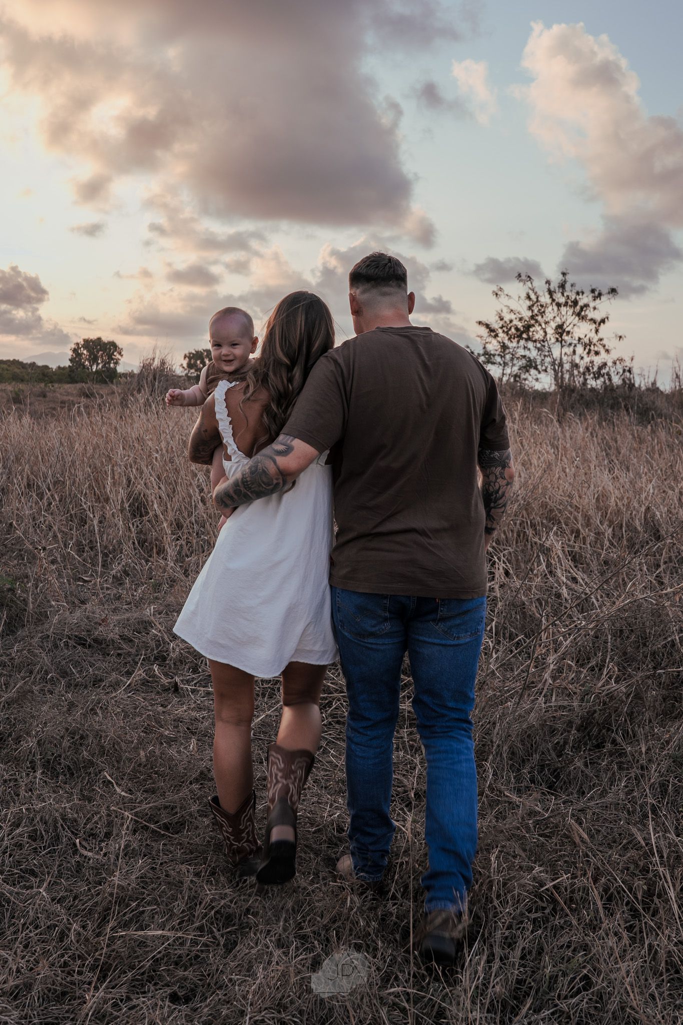 A couple embraces while walking through a field of dried grass and brush during sunset.