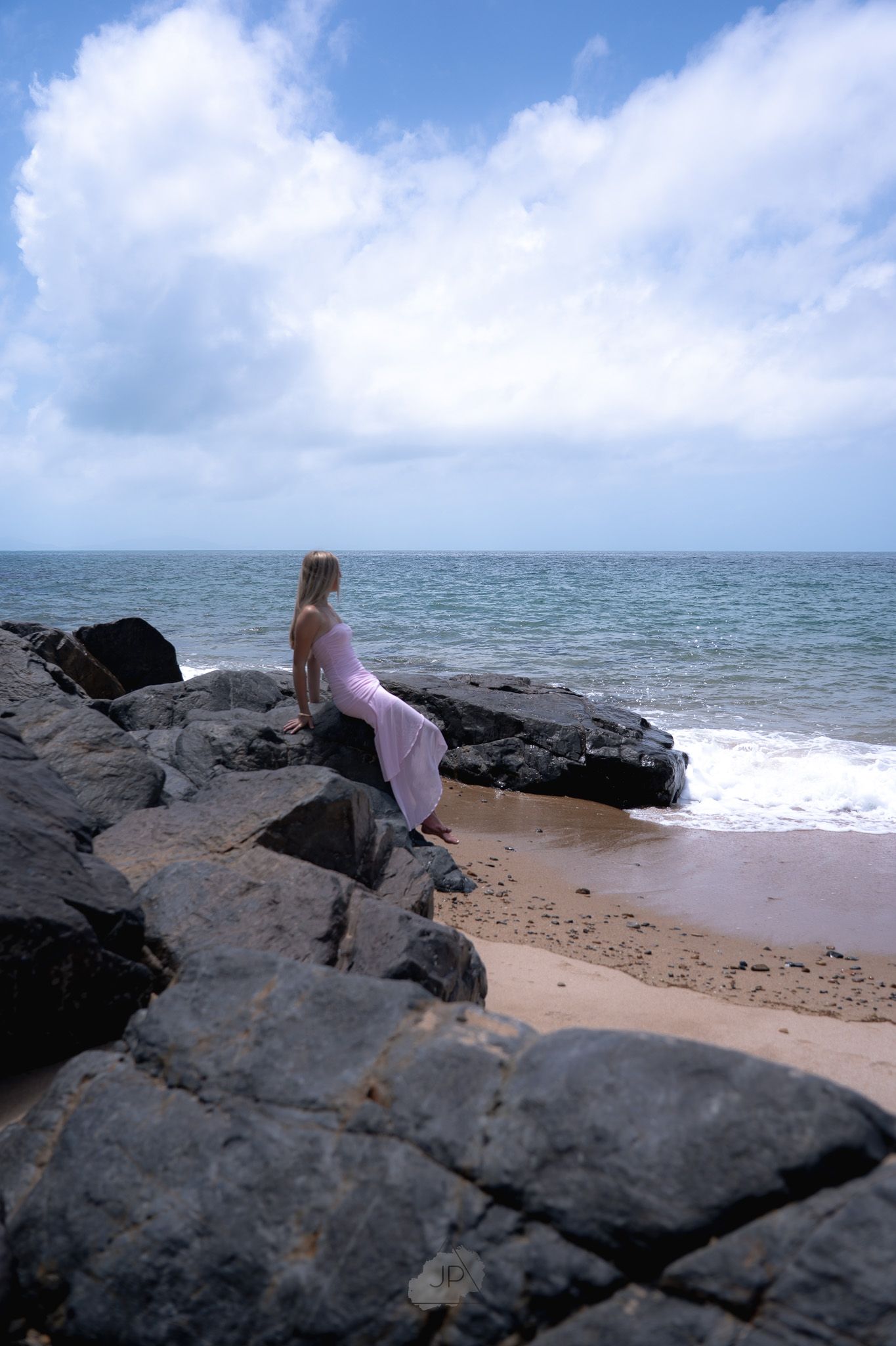 A person stands on dark volcanic rocks overlooking the ocean waves crashing against a sandy beach on a cloudy day.