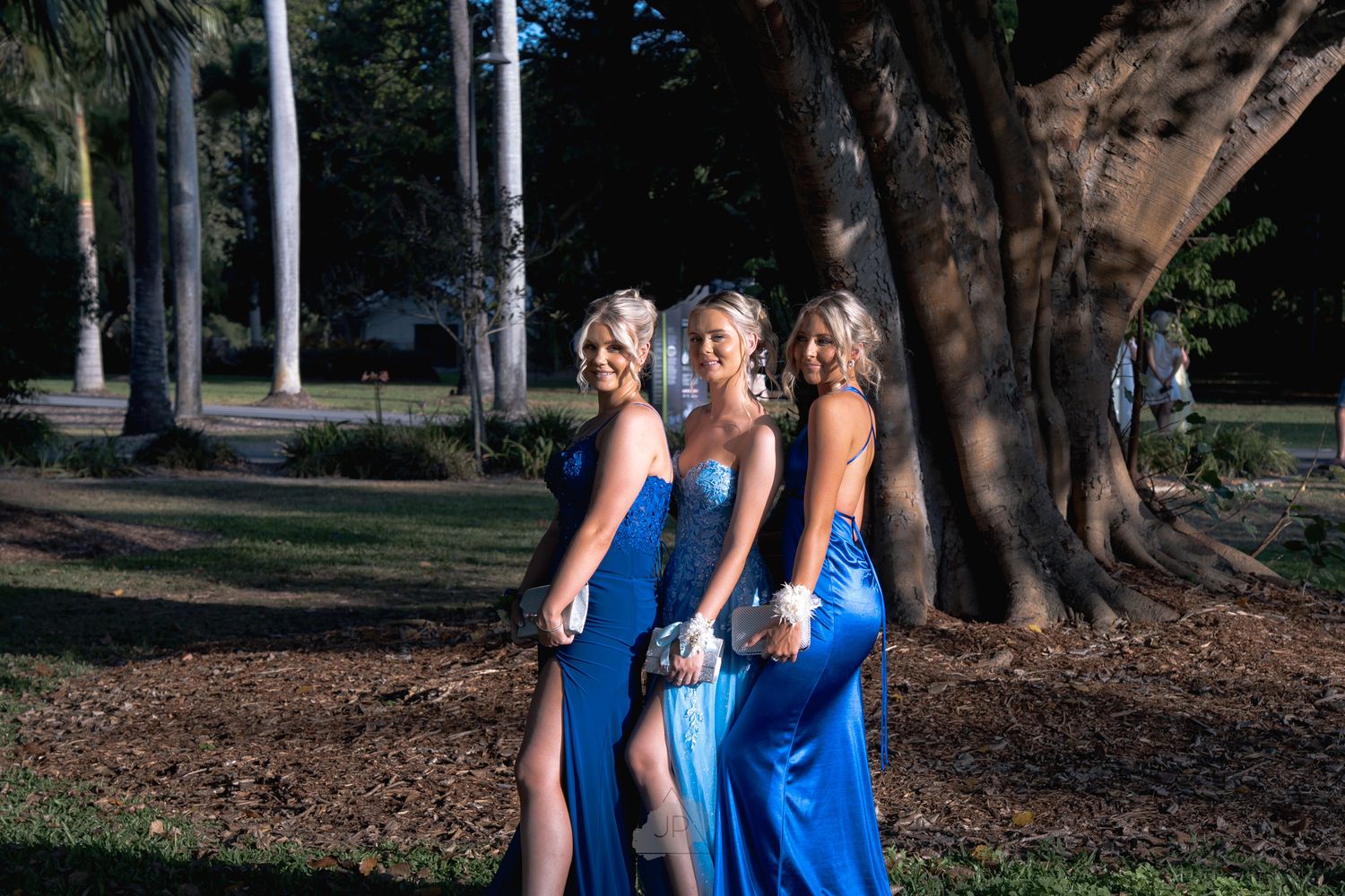 Three women in matching blue dresses pose together under a large tree in an outdoor garden setting.