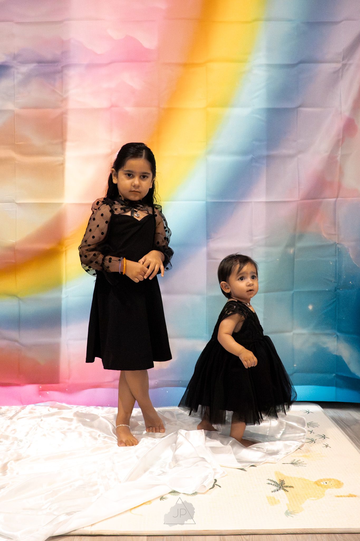 A child in a black lace dress poses against a dreamy rainbow backdrop with fluffy white clouds.