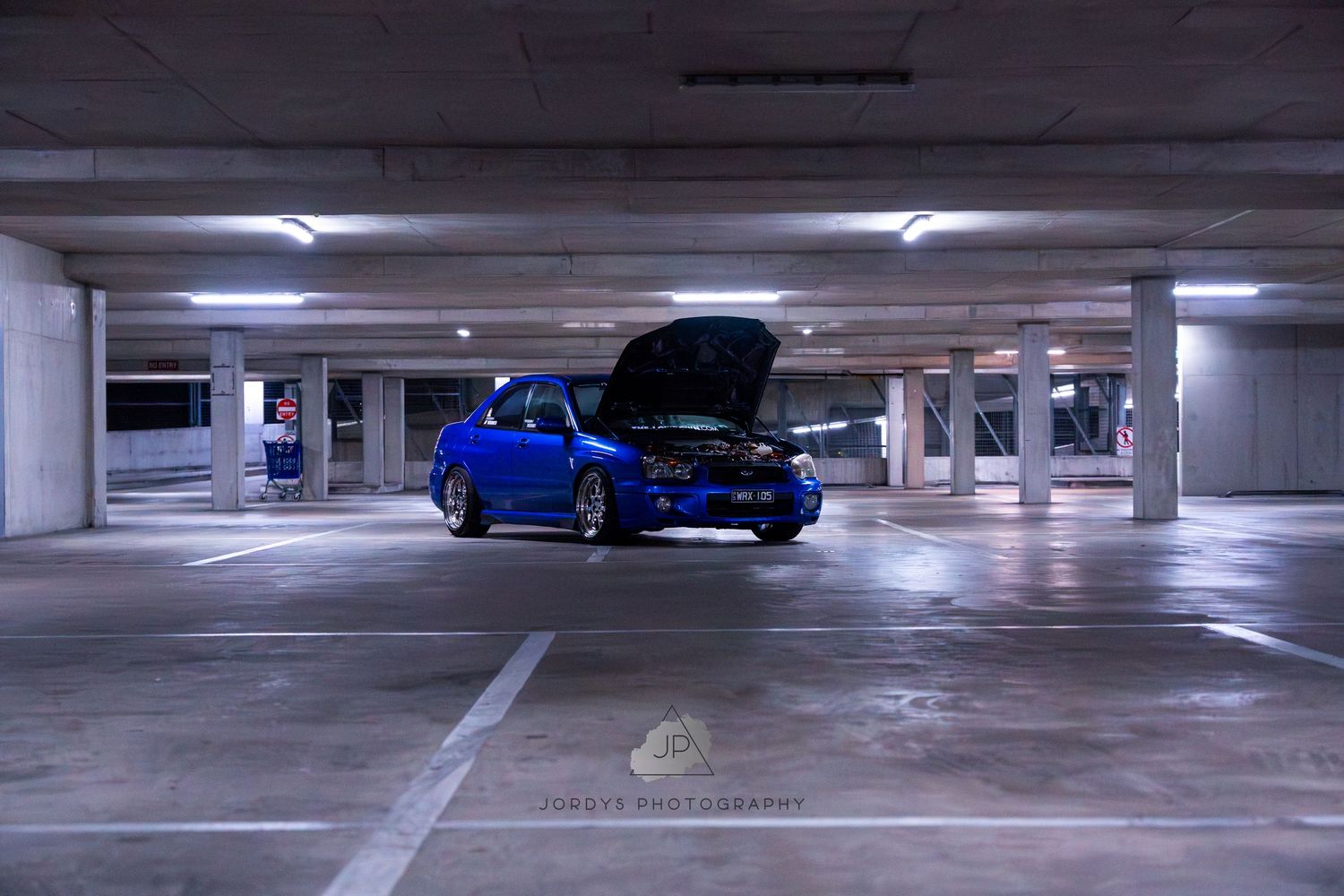A bright blue Subaru WRX with its hood up sits in a dimly lit parking garage.