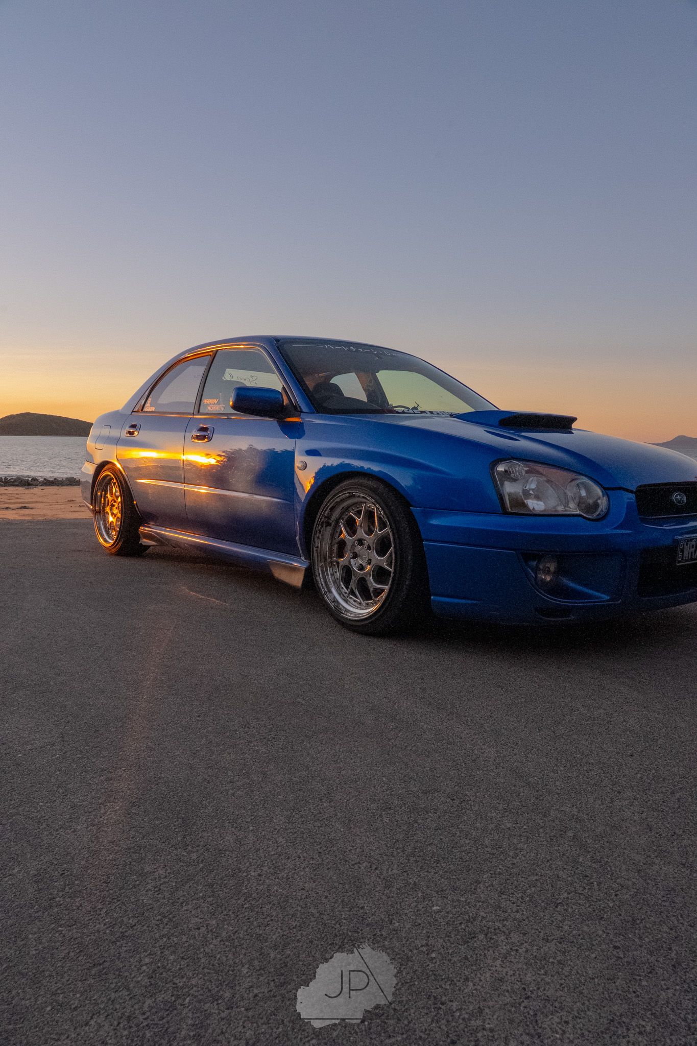 A blue modified Subaru WRX STI sits on a flat surface during sunset with dramatic lighting.