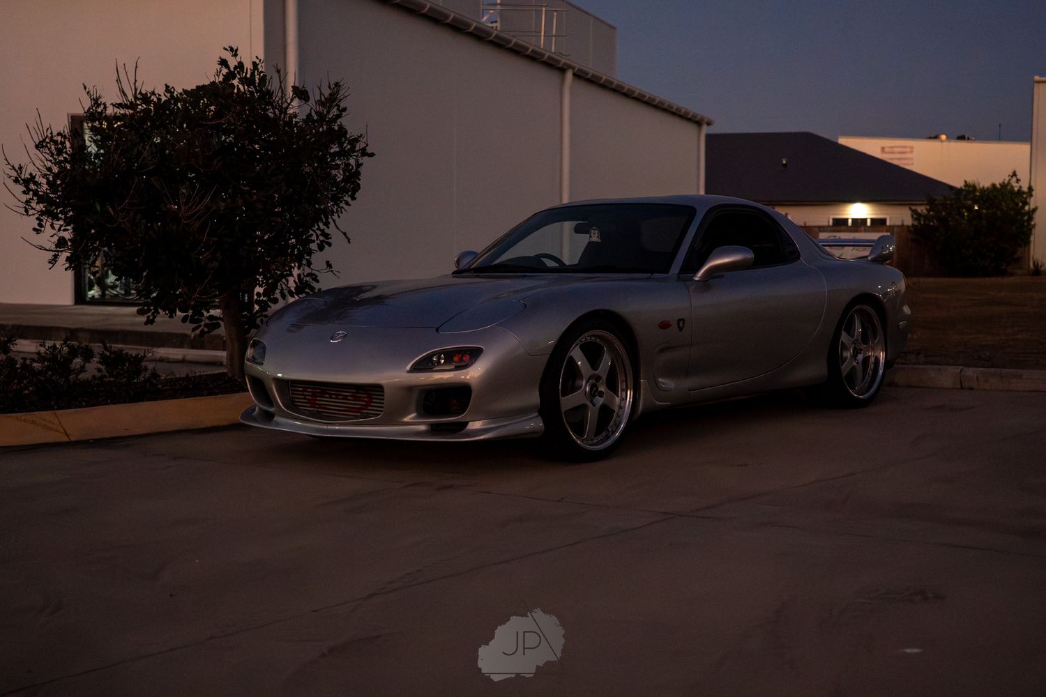 A silver sports car parked in a parking lot at twilight with trees and buildings visible in the background.