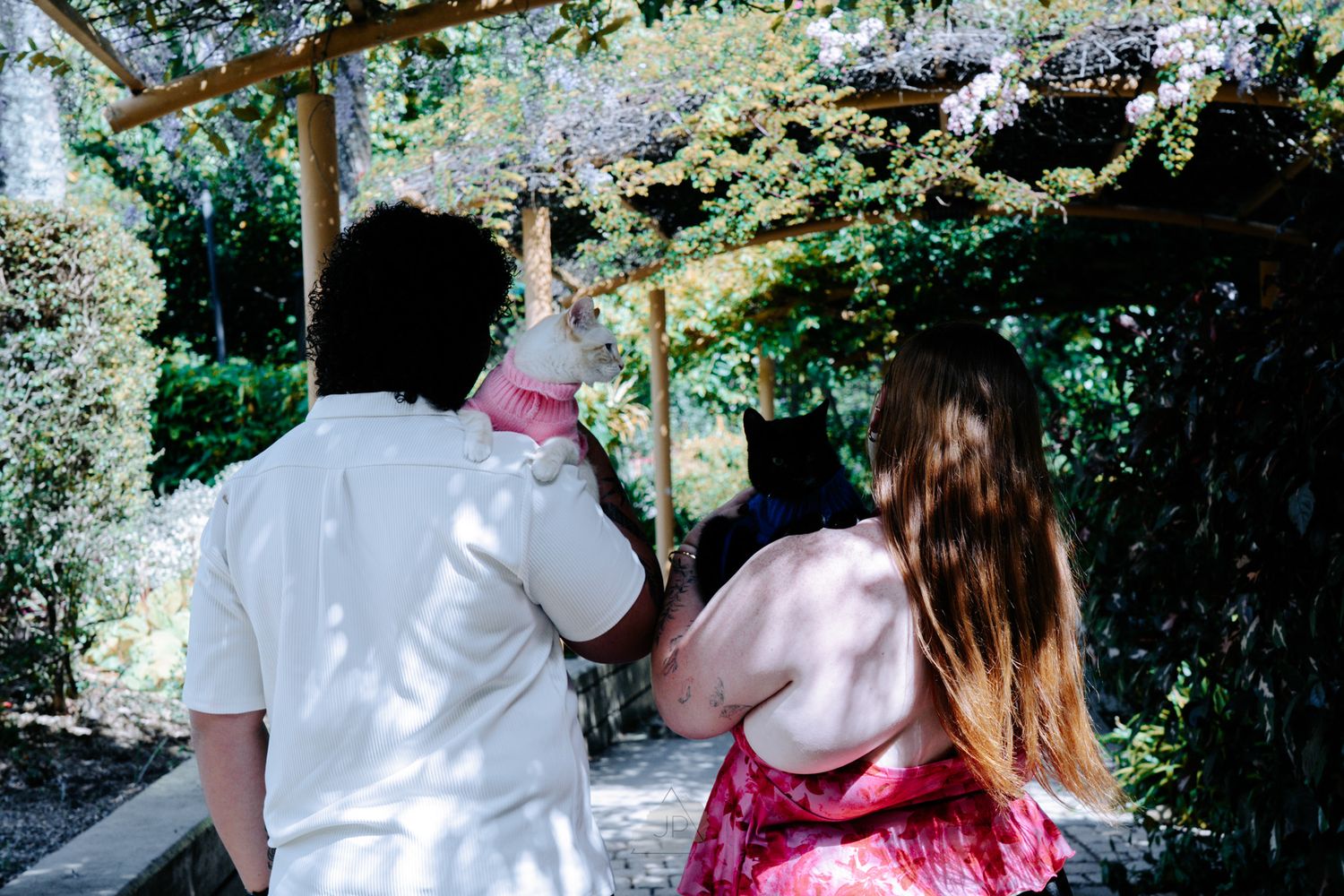 A couple in white and pink outfits walk together through a lush garden path on a sunny day.
