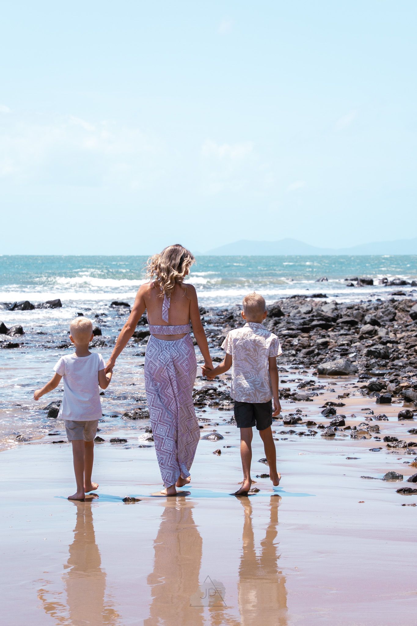 Three people in white clothing walk along a rocky beach shoreline as waves crash in the background.